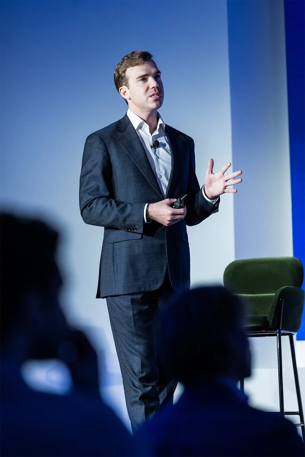 A man in a dark gray suit and white shirt is giving a presentation on stage, holding a remote control, in front of a blue background with some audience members visible in the foreground.