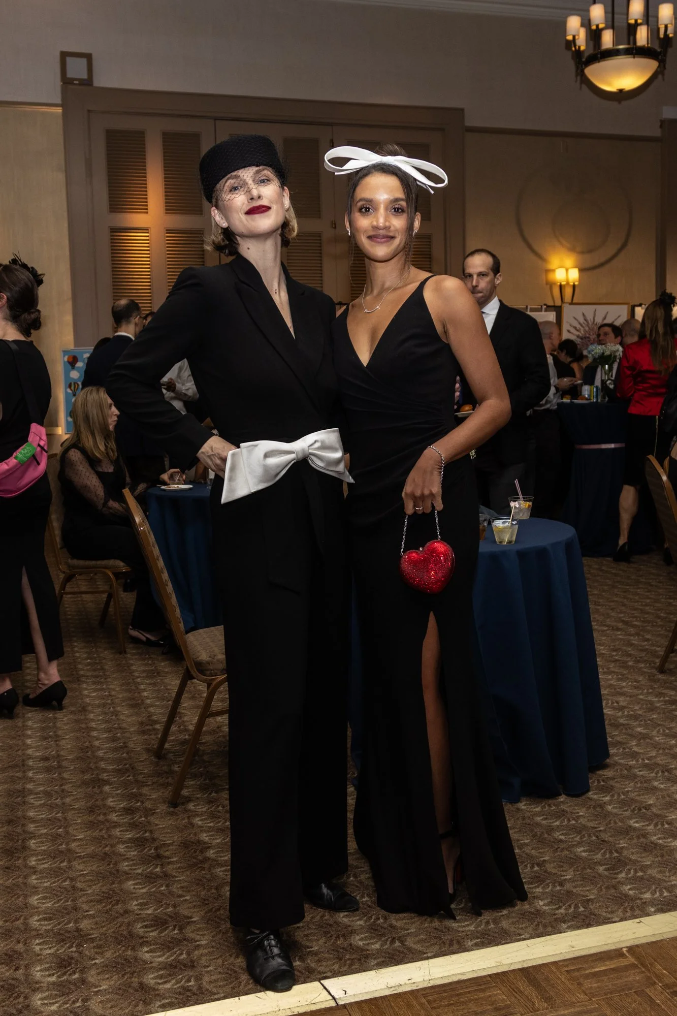 Two women pose together at a formal event, one wearing a black pantsuit with a white bow belt and black hat with netting, the other in a black dress with a white bow headpiece and red heart-shaped handbag. Several people are visible in the background