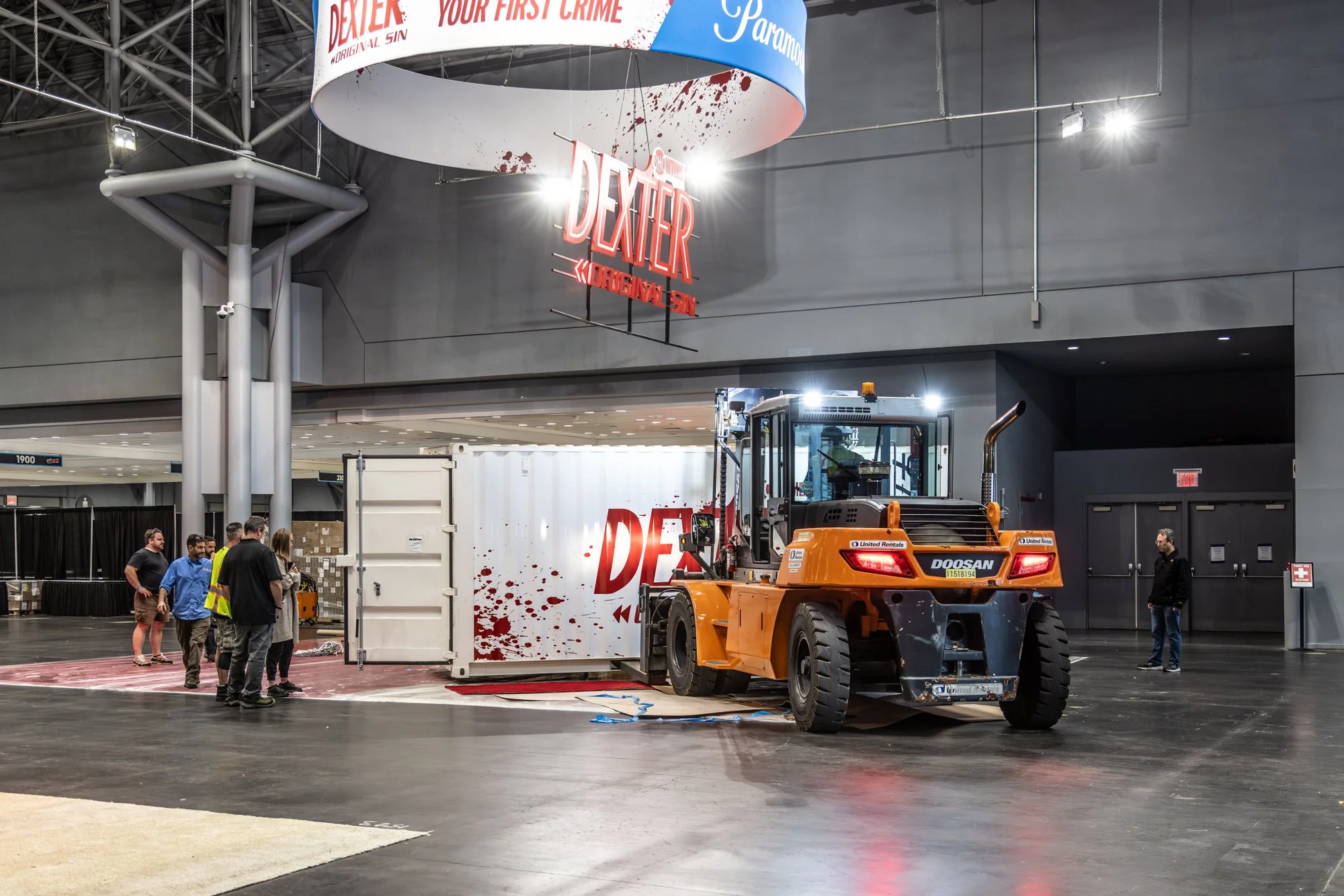 Orange Doosan forklift unloading a Dexter promotional display inside a convention center, with Paramount signage overhead