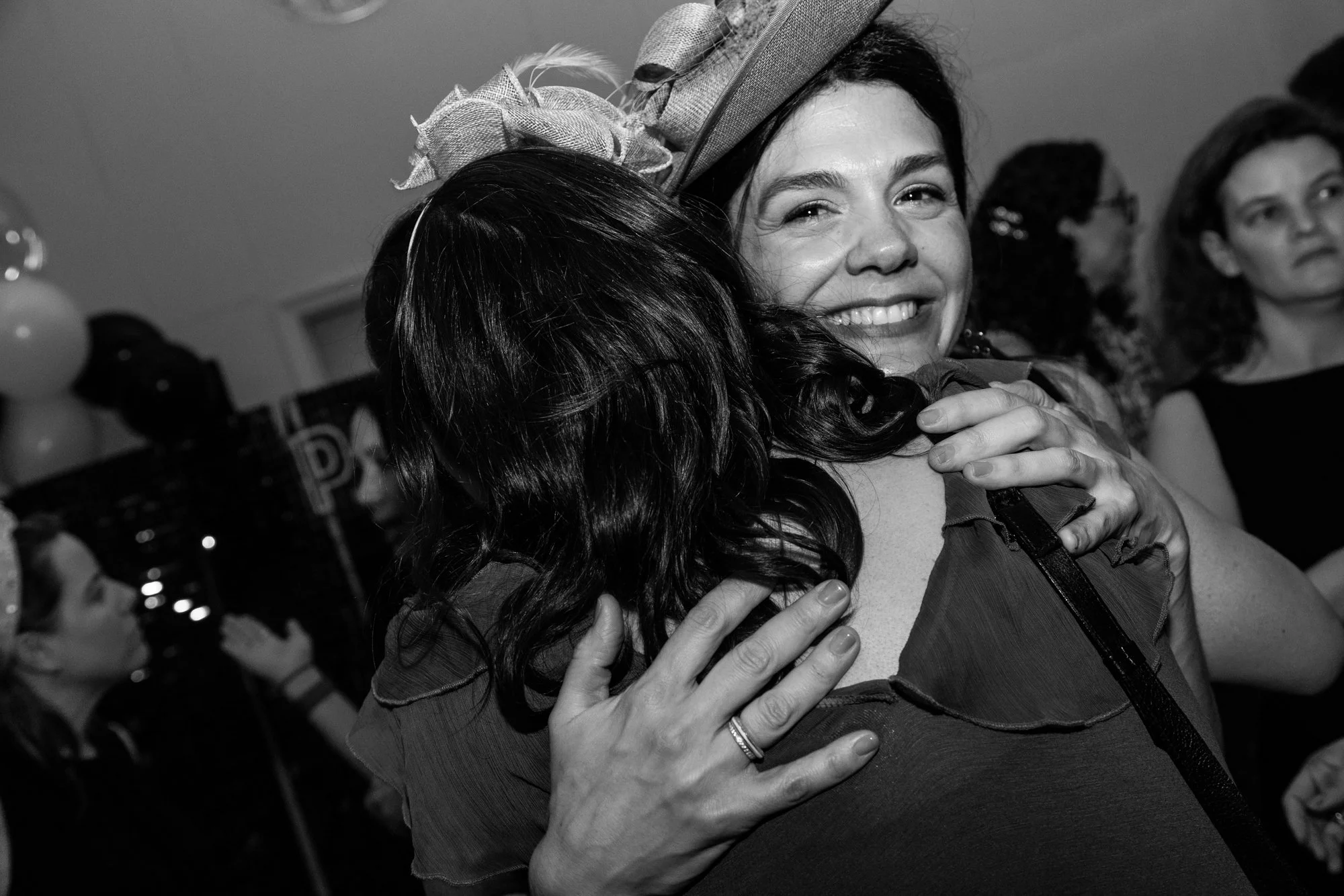 Two women hugging tightly and smiling at a celebration or party, with other people in the background.