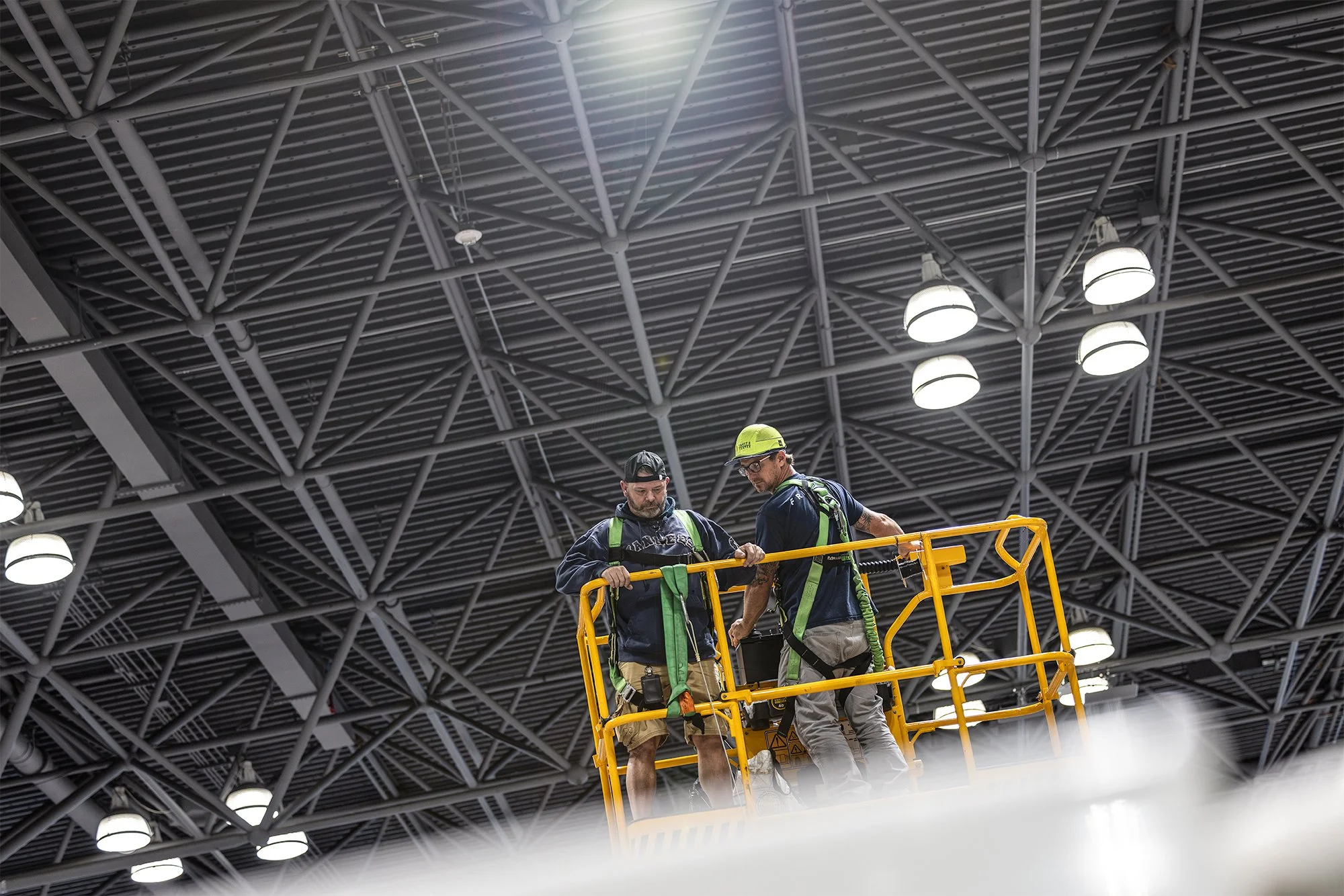 Two construction workers in safety gear stand in a yellow aerial lift beneath an industrial metal-truss ceiling.