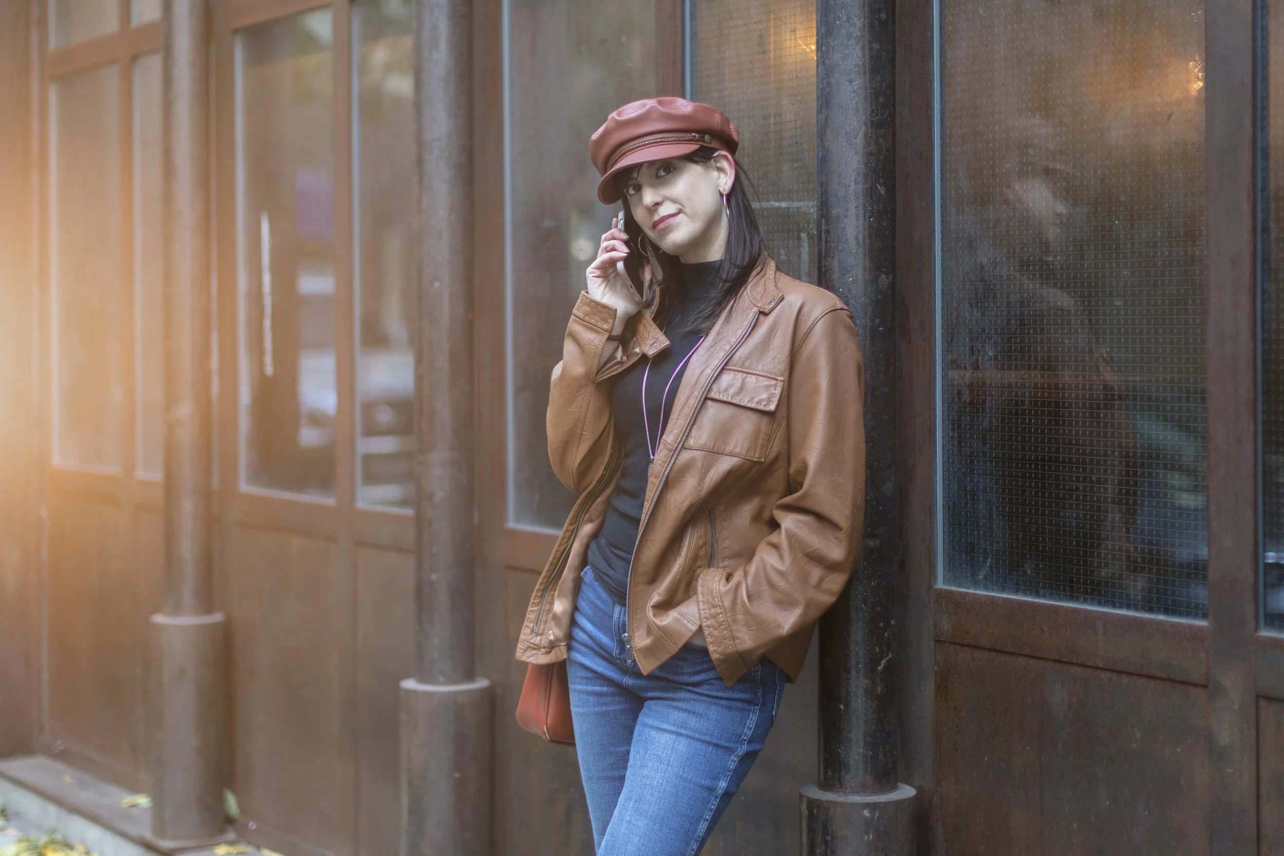 A woman with dark hair wearing a brown leather jacket, jeans, and a brown hat, standing against a wooden wall while talking on a cellphone.