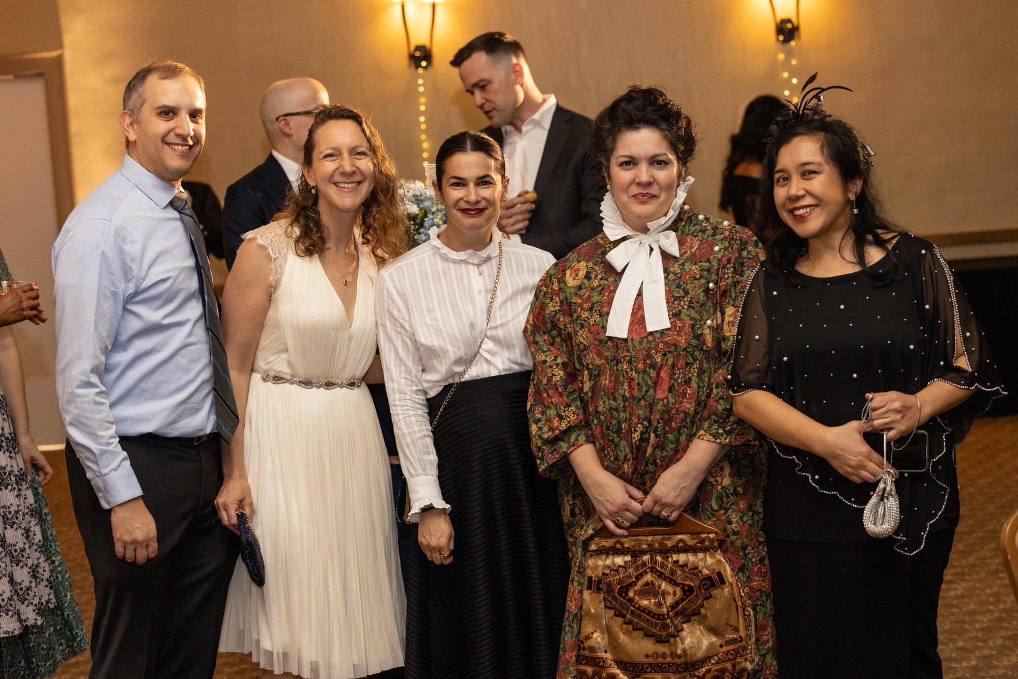 Group of six diverse adults dressed in formal attire at an indoor event with warm lighting and decorative wall lamps.
