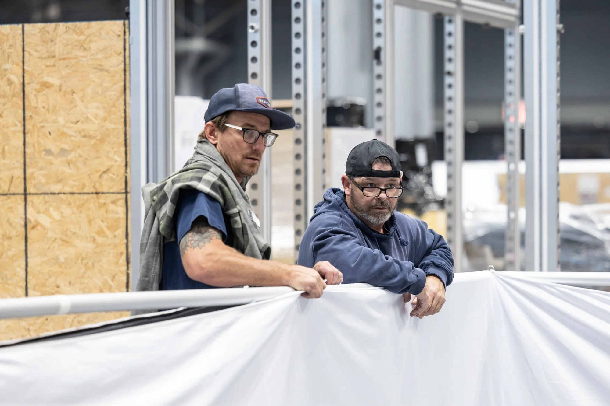 Two workers wearing caps and glasses handle a large sheet of white material inside an industrial workshop.