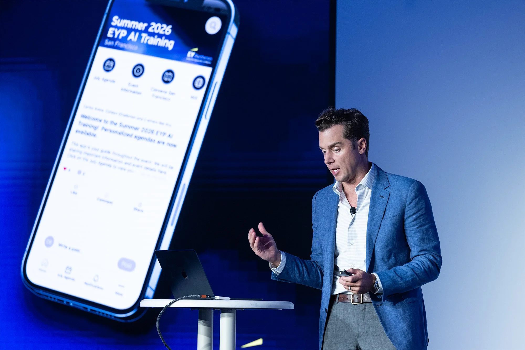 A man in a blue blazer giving a presentation with a large digital screen displaying a phone screen with text about a summer training event.