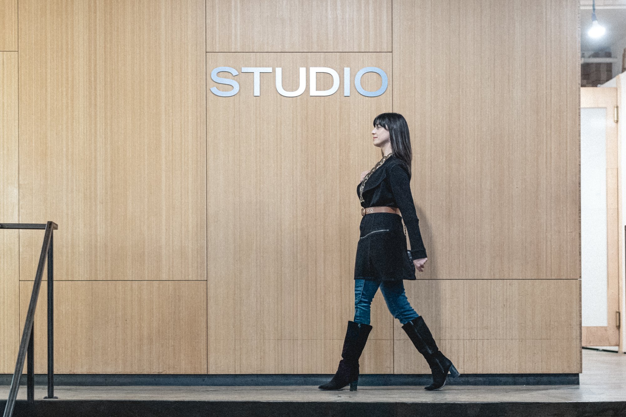 A woman walking past a wall with the sign 'STUDIO' in large letters, wearing a black outfit and high-heeled boots.