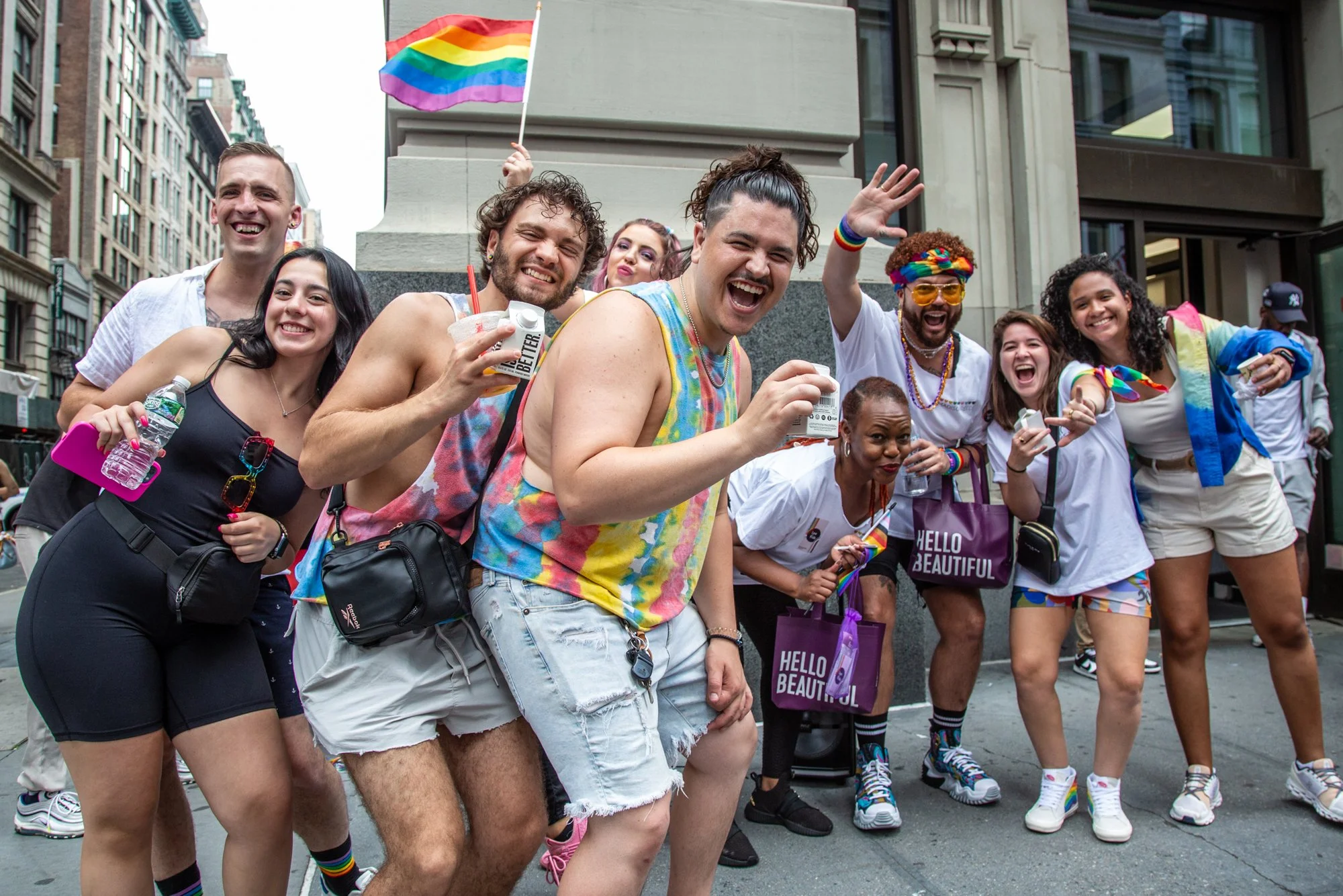 Smiling group celebrating Pride in a city street with rainbow flags, colorful clothing, and accessories.