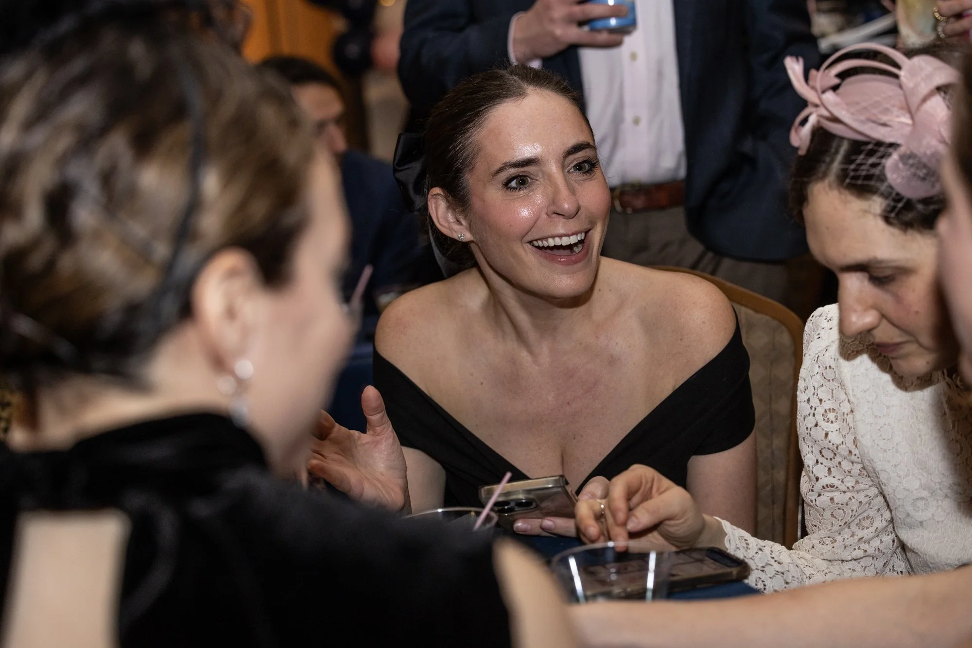 A group of women are gathered around a table at a social event. One woman in the center, with dark hair and wearing a black dress, is smiling and engaging with others. The woman to her right, wearing a white lace dress and pink fascinator, is looking