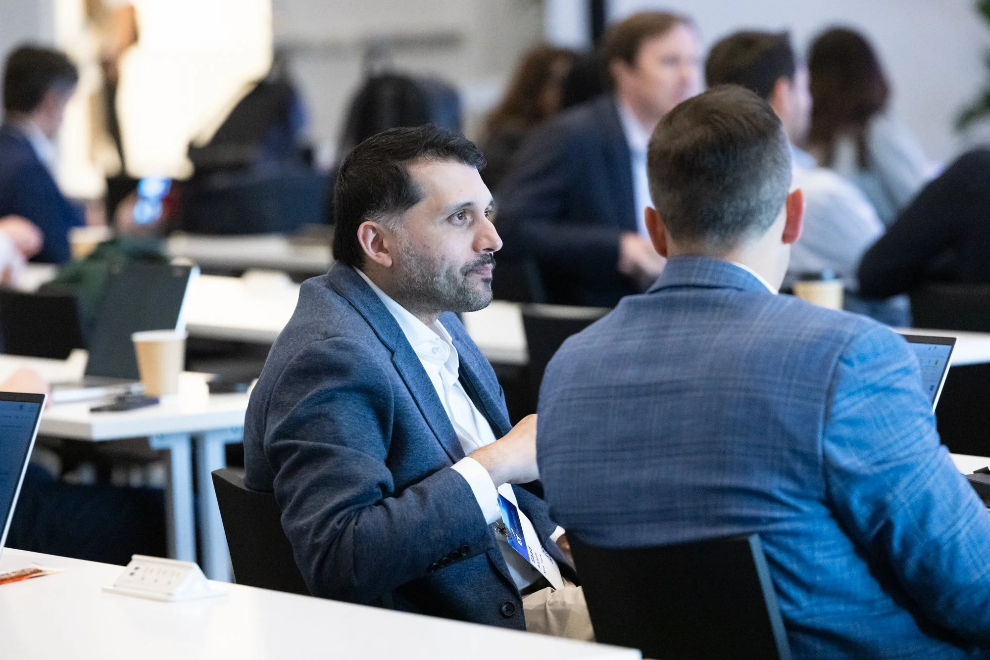 A group of professionals in business attire attending a conference or seminar, sitting at tables with laptops and documents, listening attentively.