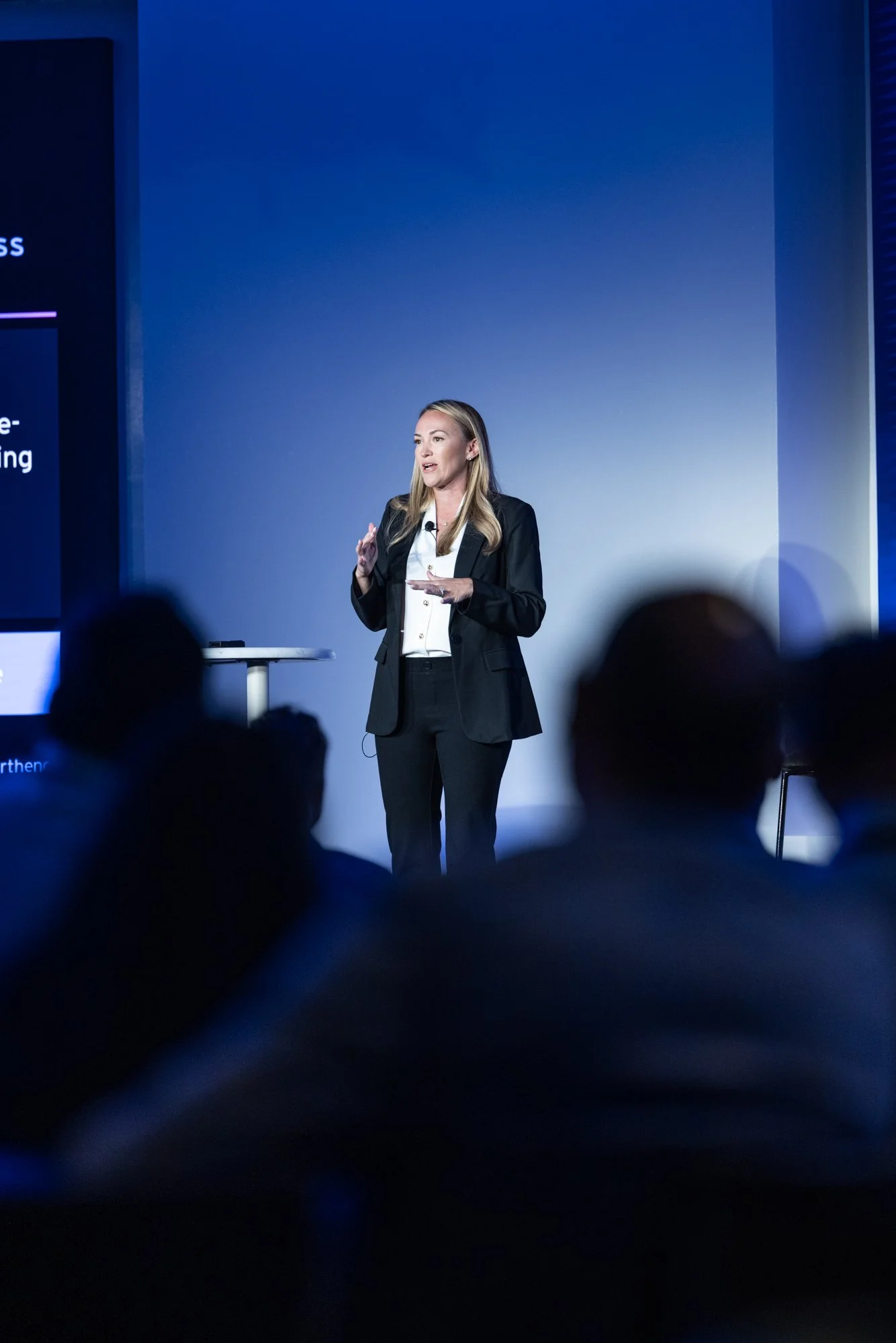 A woman in a black suit giving a presentation on stage. She is speaking and gesturing with her right hand, and there is a microphone attached to her lapel. The background is blue with presentation slides partially visible.