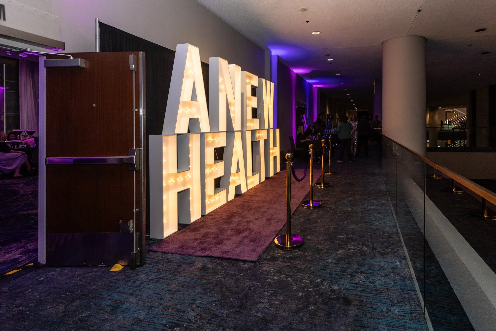 Lit marquee sign spelling out 'ANNIVERSARY' at an event, with people socializing nearby in a venue with dark flooring and purple lighting.