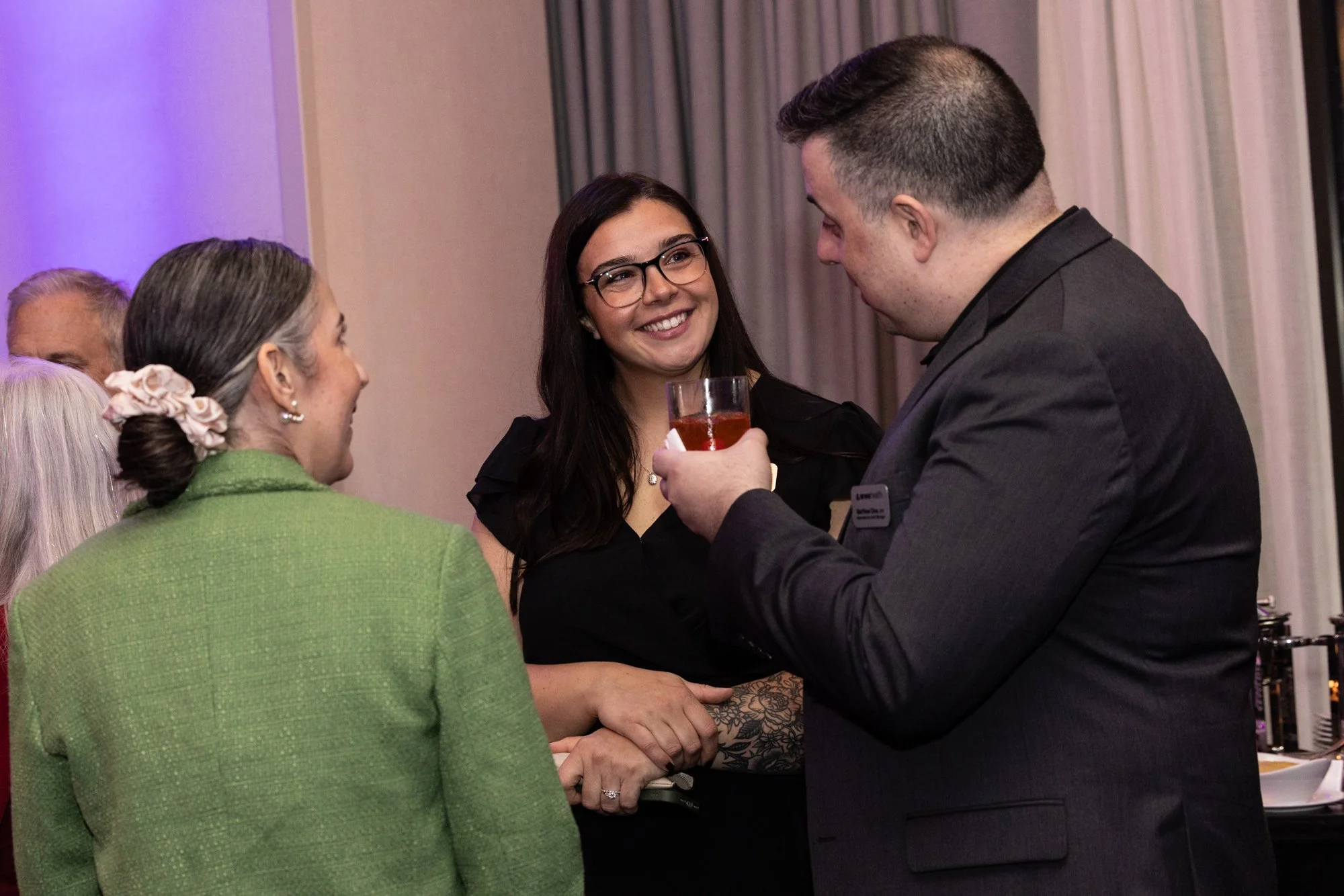 Group of people at a social gathering, including a woman with dark hair and glasses smiling, a woman with gray hair in a green jacket with a flower hair clip, and a man with short dark hair in a suit holding a drink, engaged in conversation indoors.