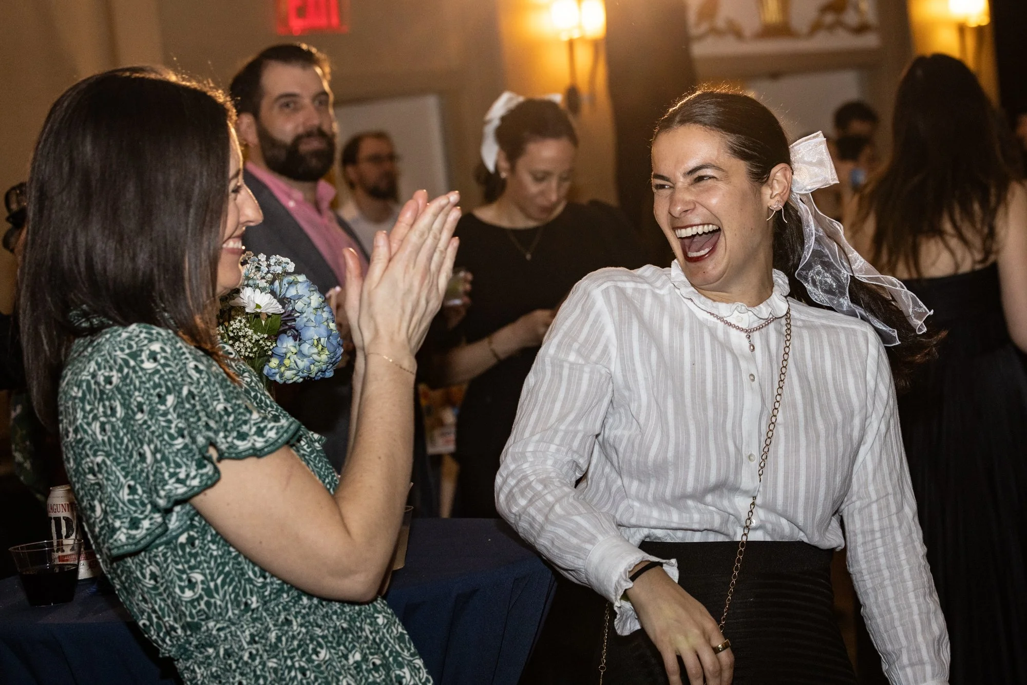 Two women are laughing and high-fiving at a celebration, with a group of people in the background, in an indoor setting with warm lighting.