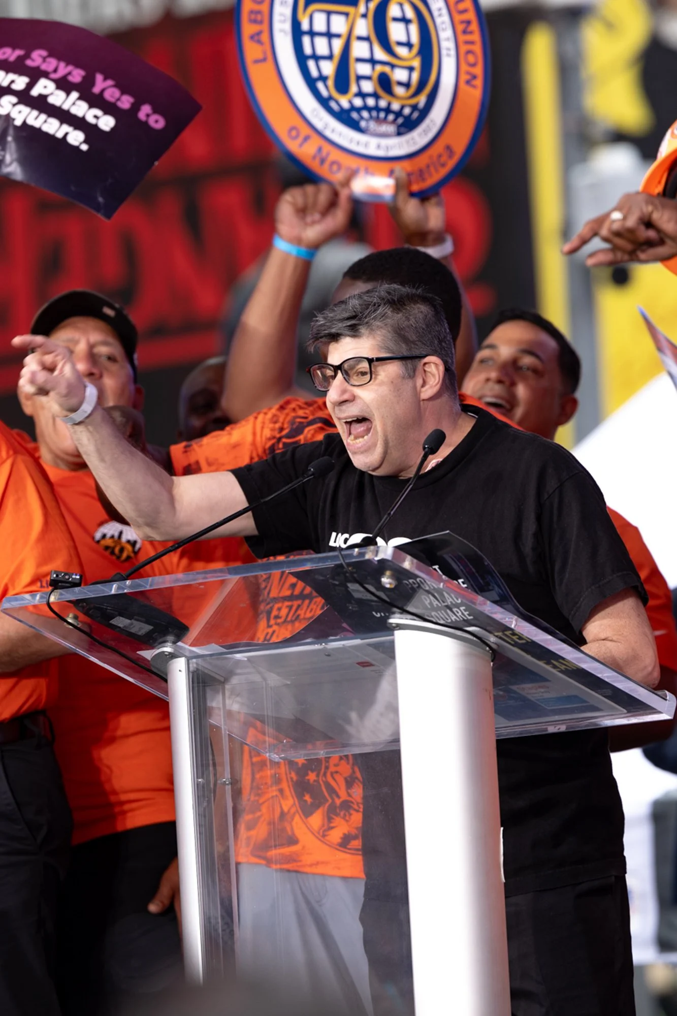 Man with glasses speaking at a podium during a rally, surrounded by supporters holding signs and cheering.