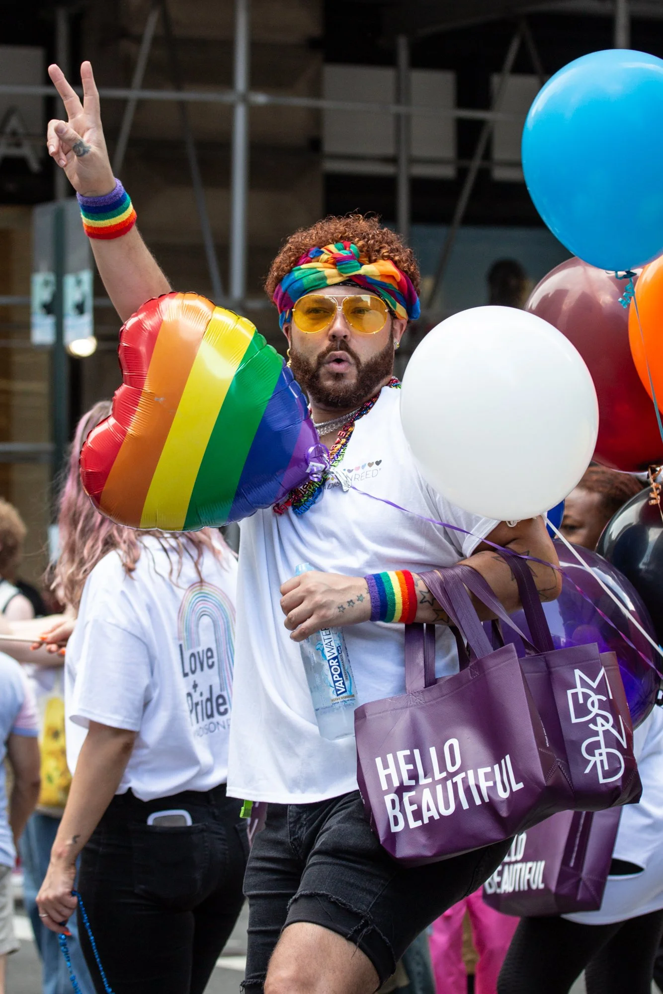 Pride parade participant wearing rainbow accessories, yellow sunglasses, and a colorful headband while holding balloons and a rainbow heart balloon.