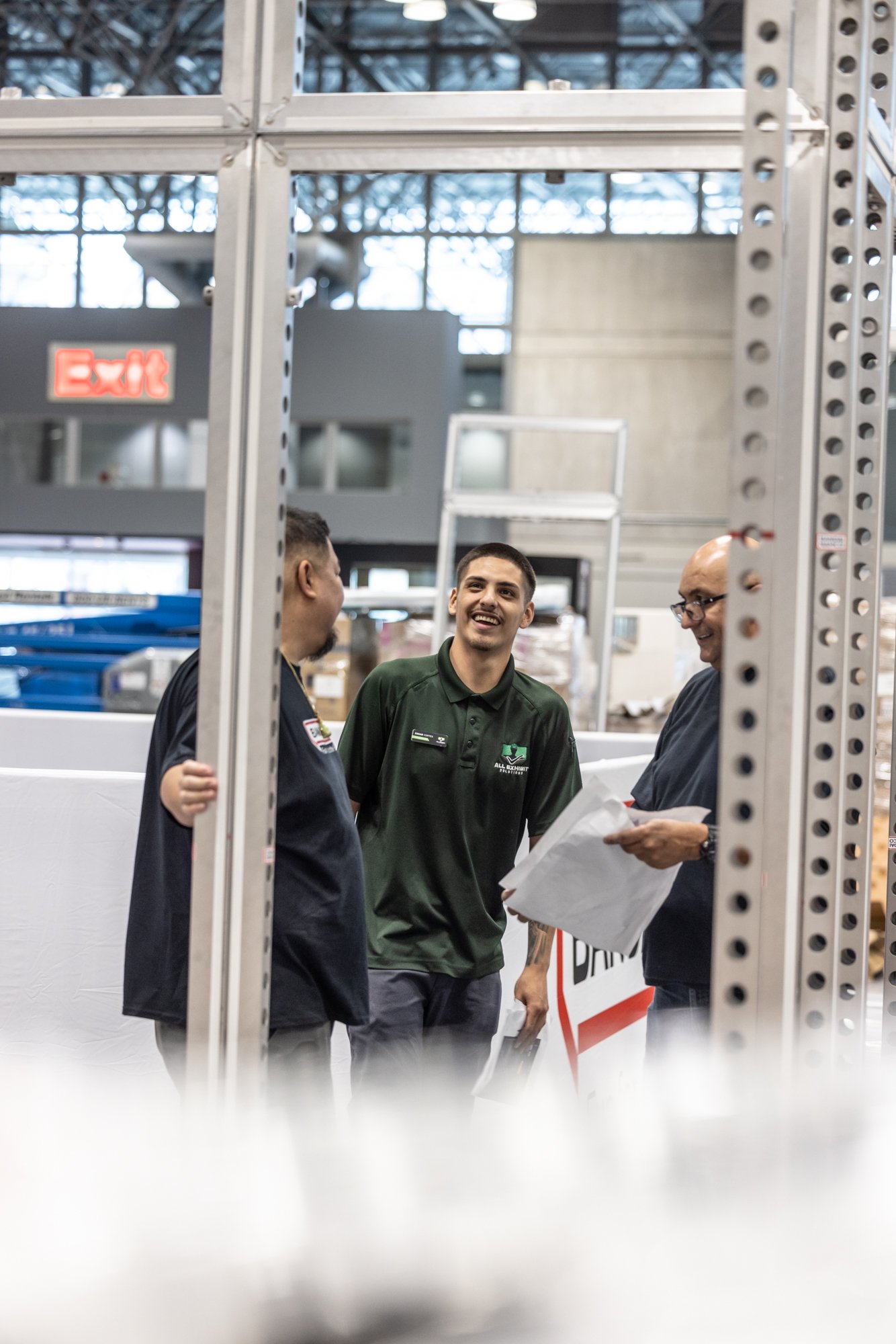 Three coworkers talking inside an industrial facility, with metal support beams and a ladder in the background.