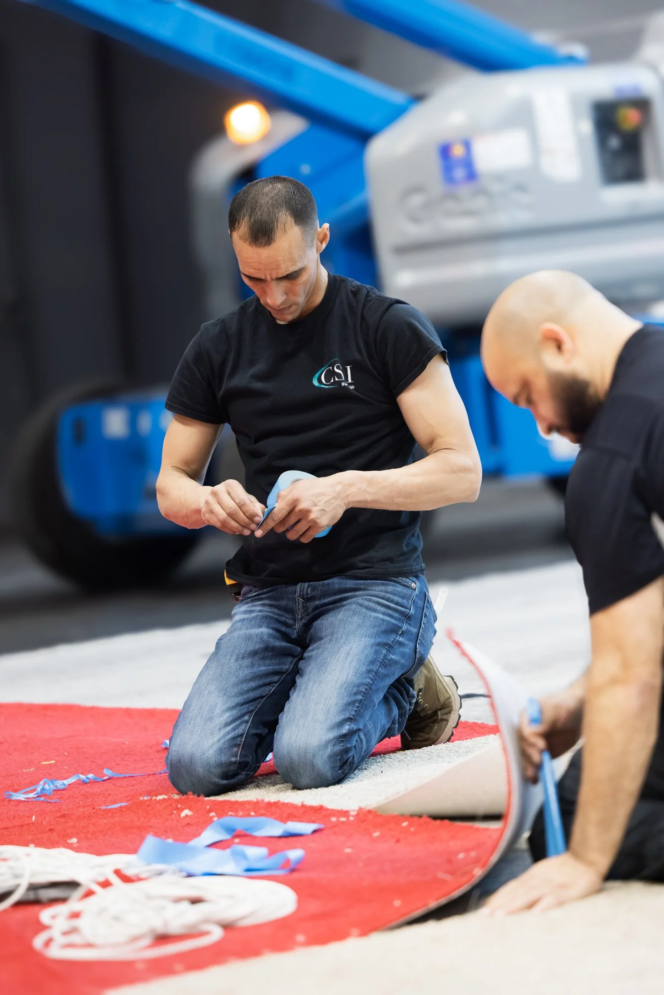 Two men kneeling on a red mat inside a gym, handling blue straps near exercise equipment.
