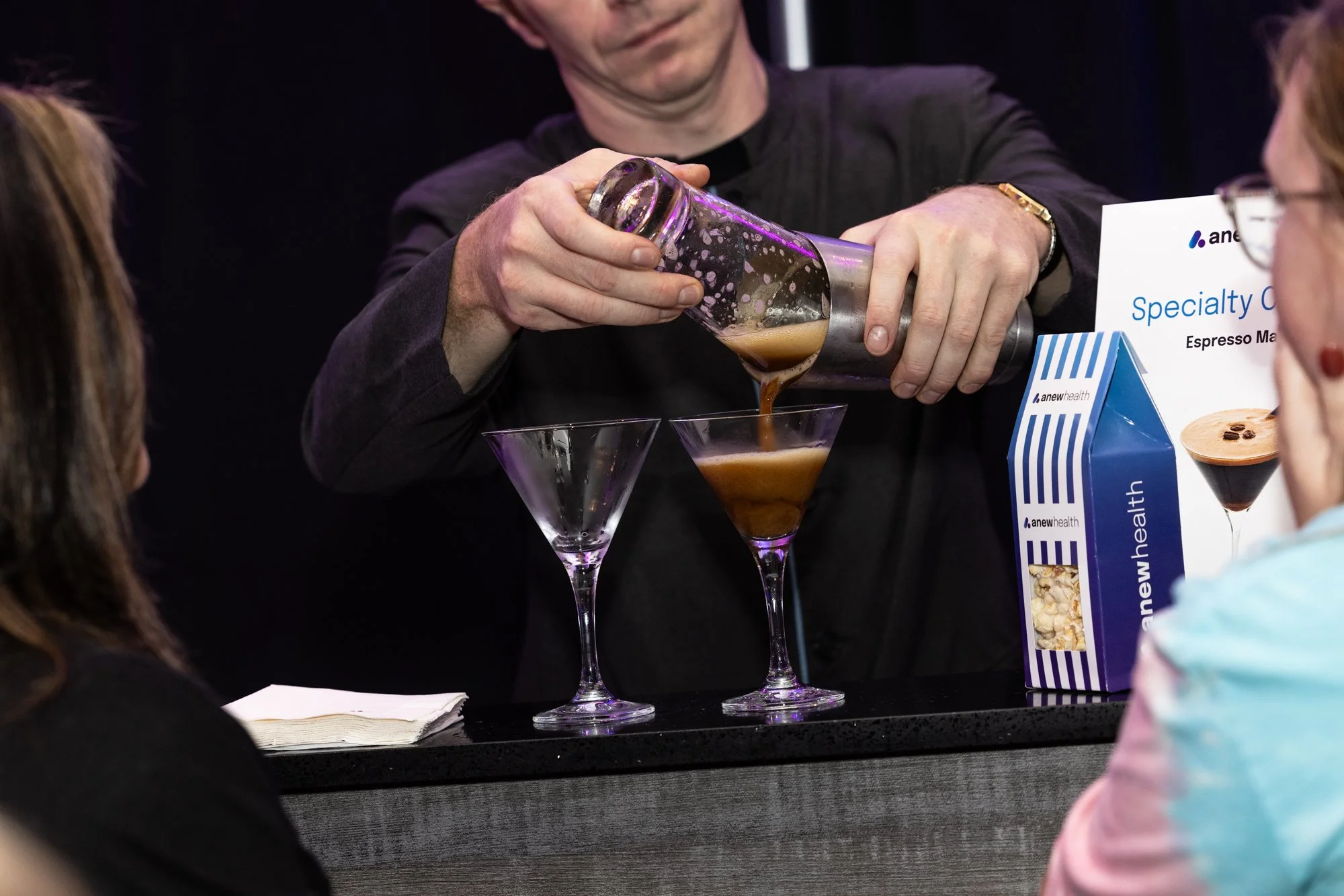A person pouring a coffee-colored beverage into a martini glass at a promotional event, with a sign featuring images of coffee and a box of coffee mix beside them, and onlookers watching.