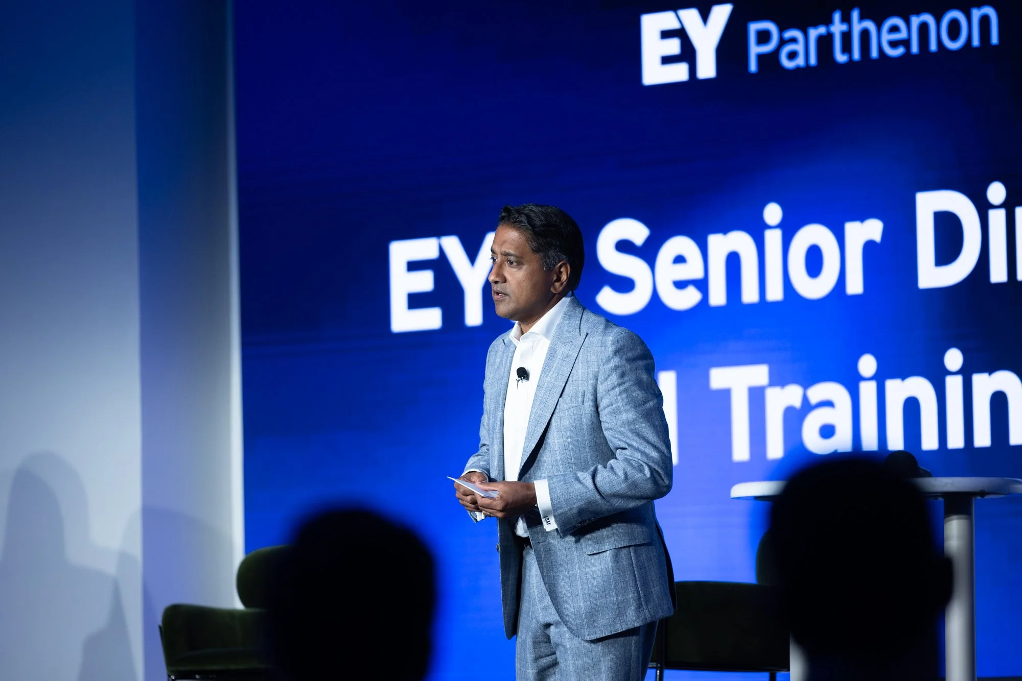 A man in a gray suit speaking at a conference with a large blue screen behind him that has white text reading "EY Parthenon" and partially visible words related to training.