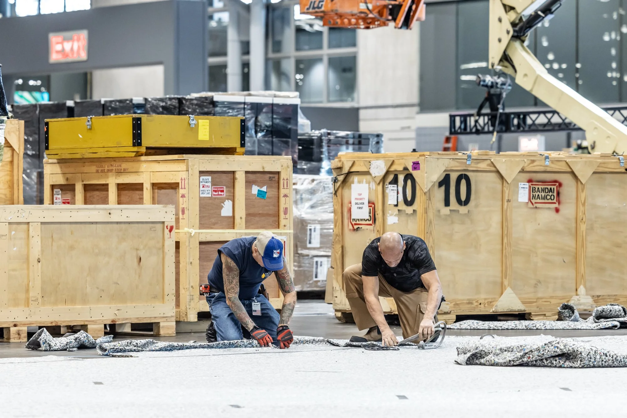 Two workers install flooring beside large wooden shipping crates in an indoor construction or event setup area.