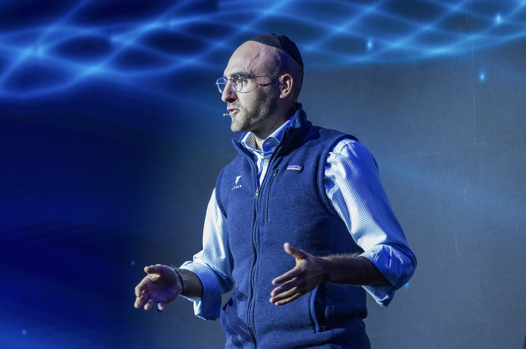 A man wearing glasses and a dark head covering delivering a presentation on stage with a blue digital background.