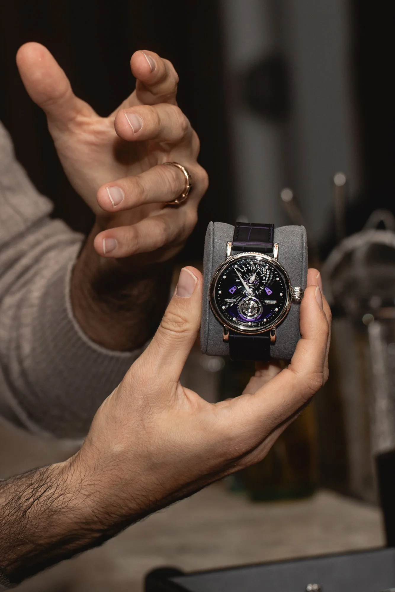 Close-up of a person holding a luxury wristwatch with a black strap and a black dial, showing detailed watch features, while another person points towards it.