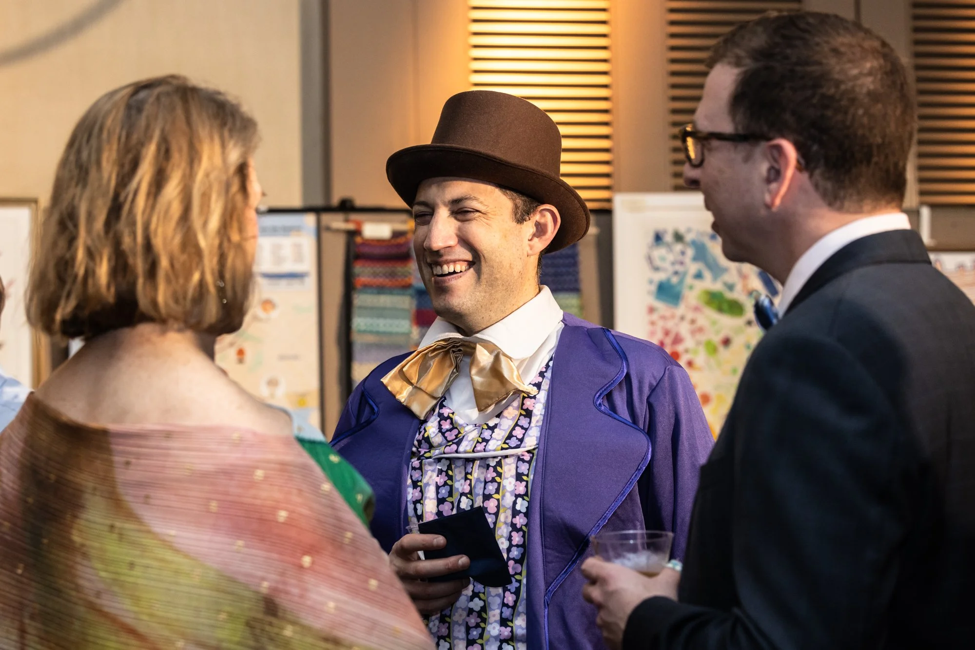 Three people are talking at a social gathering. One woman has shoulder-length blonde hair and is wearing a colorful, shiny dress. The man in the center is smiling, wearing a brown hat, purple jacket, and floral shirt. Another man on the right is wear