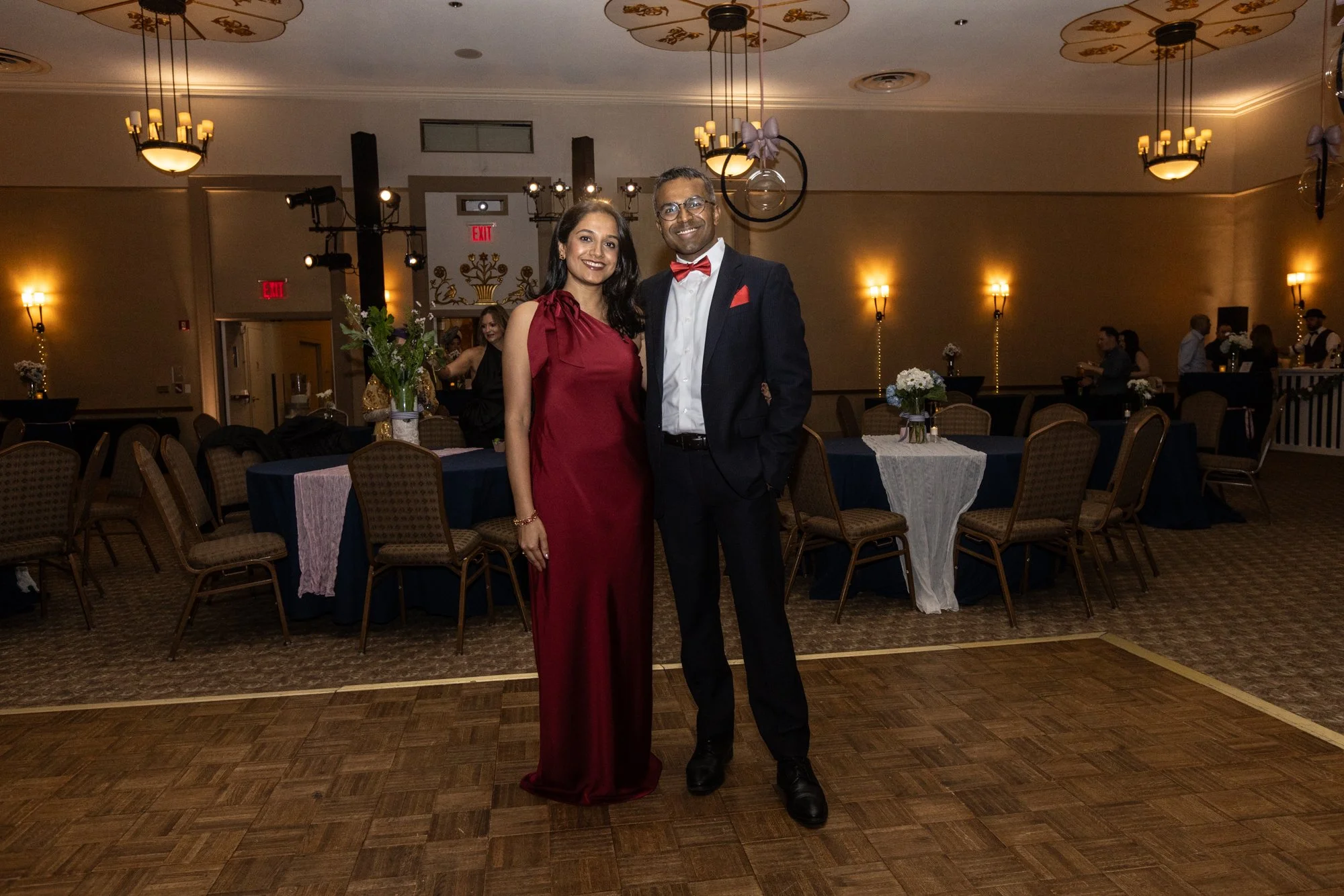A couple dressed in formal attire posing in a decorated event hall. The woman wears a sleeveless, burgundy gown, and the man wears a black suit with a white shirt and red bow tie. The hall has round tables with floral centerpieces, and there are peop