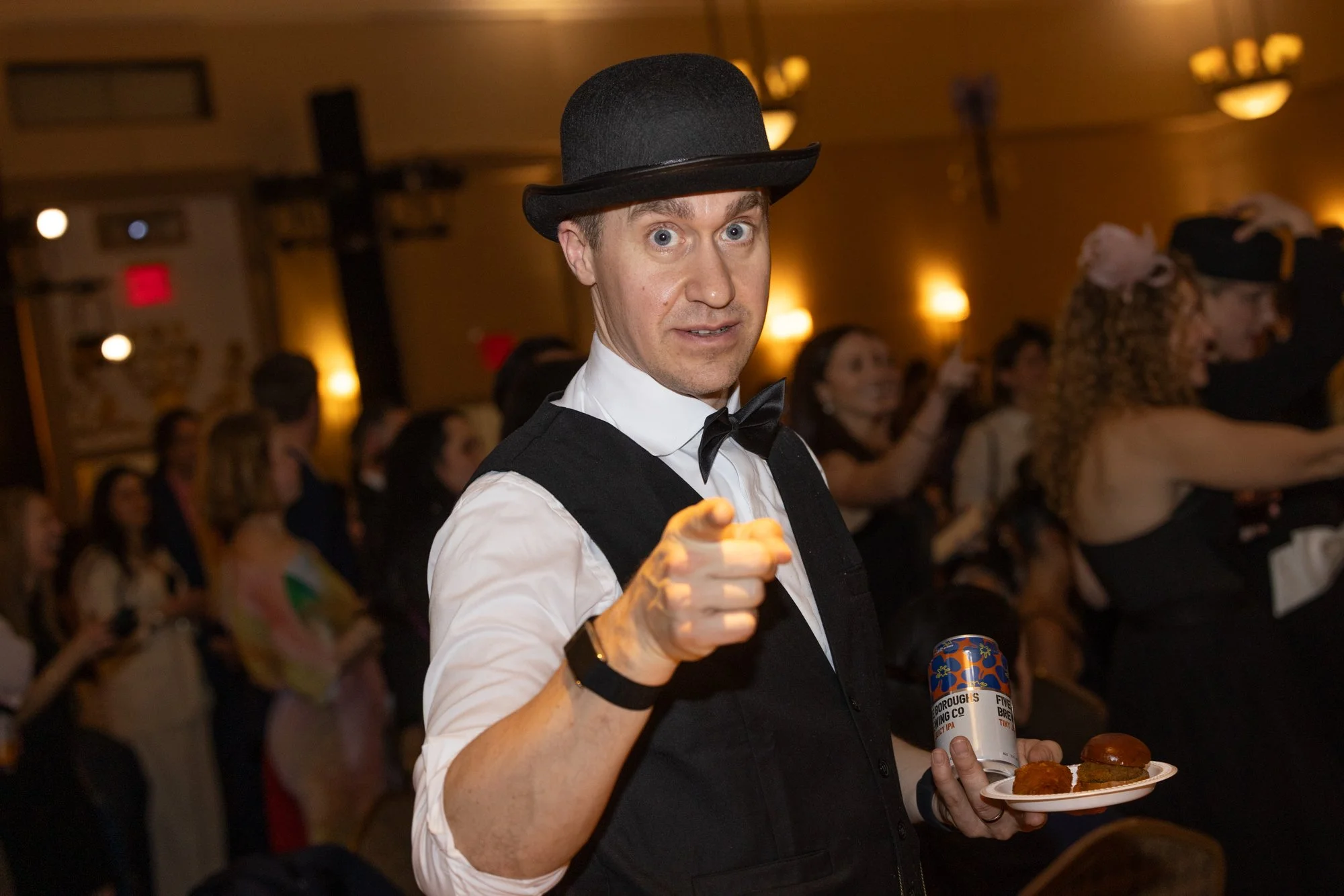 A man dressed as a waiter at a festive event, wearing a black bowler hat, white shirt, black vest, and black bow tie. He is holding a plate with mini hamburgers and pointing towards the camera with a playful expression. In the background, there are m