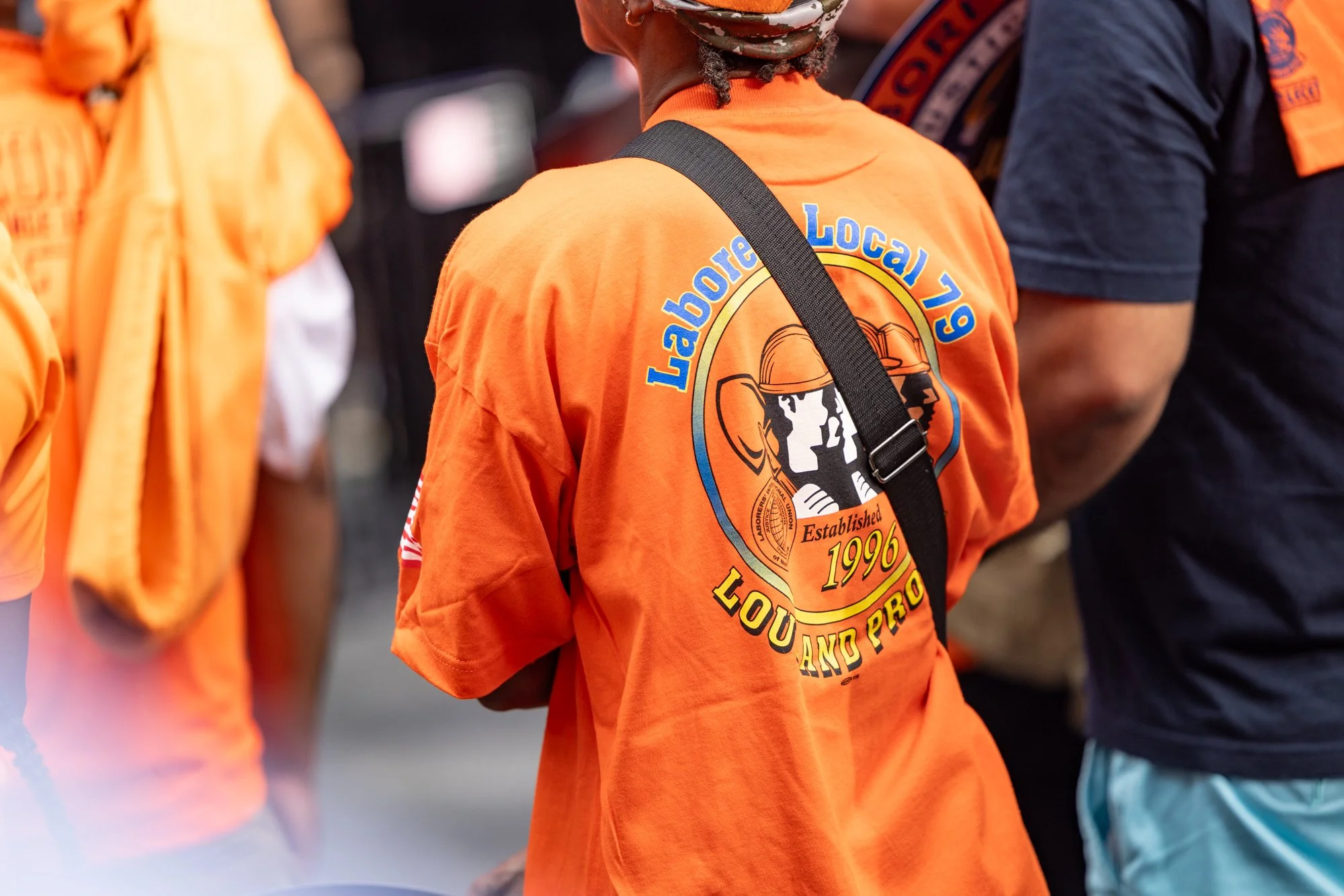 Close-up of a person wearing an orange T-shirt with the logo of Laborers' Local 79 Union, featuring a hard hat and the text 'Established 1996'. The person is carrying a black strap over their shoulder, and there are blurred people around wearing simi