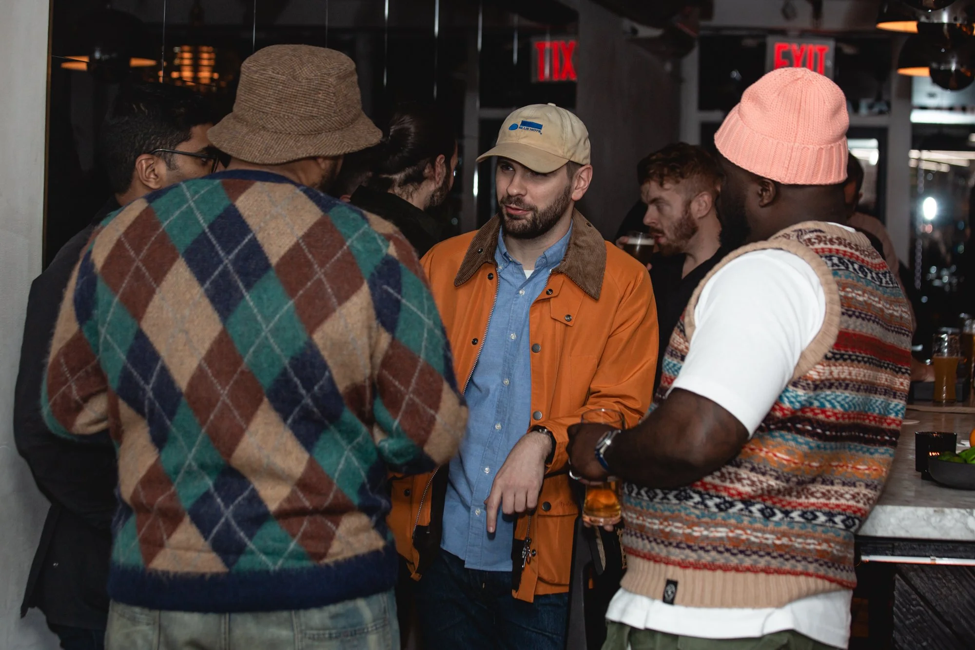 A group of six men gathered and talking closely in a bar or pub. One man in a tan cap and orange jacket appears to be speaking, while others listen. The men are wearing casual clothing with various hats and patterns. The background shows exit signs a