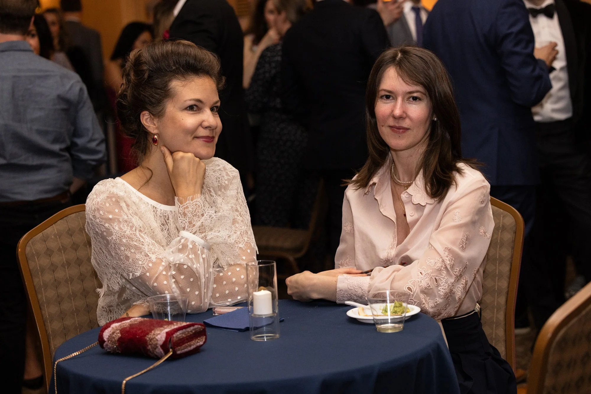 Two women sitting at a banquet table during a formal event. The woman on the left has curly brown hair and is wearing a white lace dress, while the woman on the right has straight brown hair and is wearing a light pink embroidered blouse. They are lo