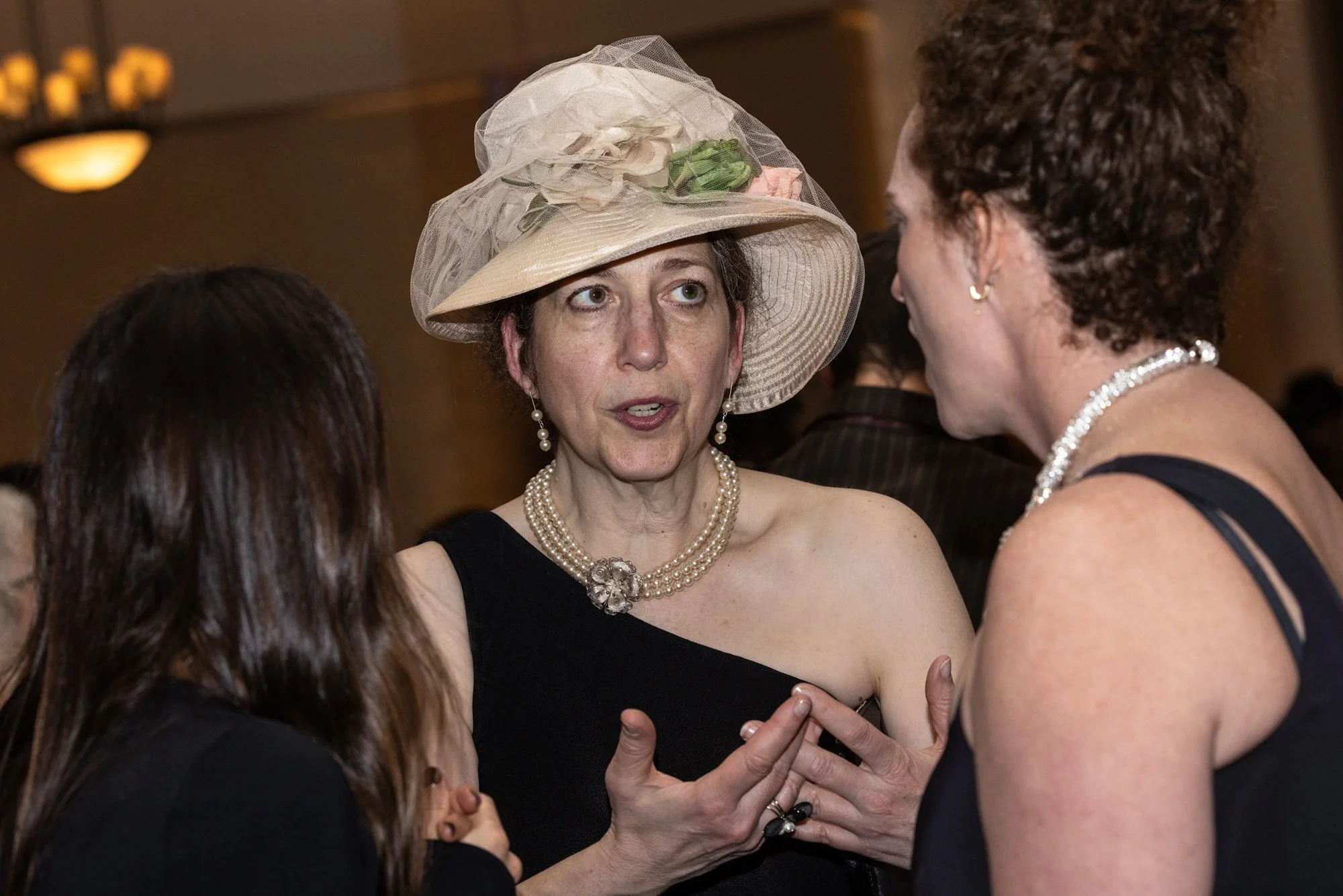 Three women wearing black dresses engaged in conversation at an indoor social event. The woman in the center wears a large beige hat with decorative netting and flowers, a pearl necklace, pearl earrings, and a brooch. The woman on the right has curly