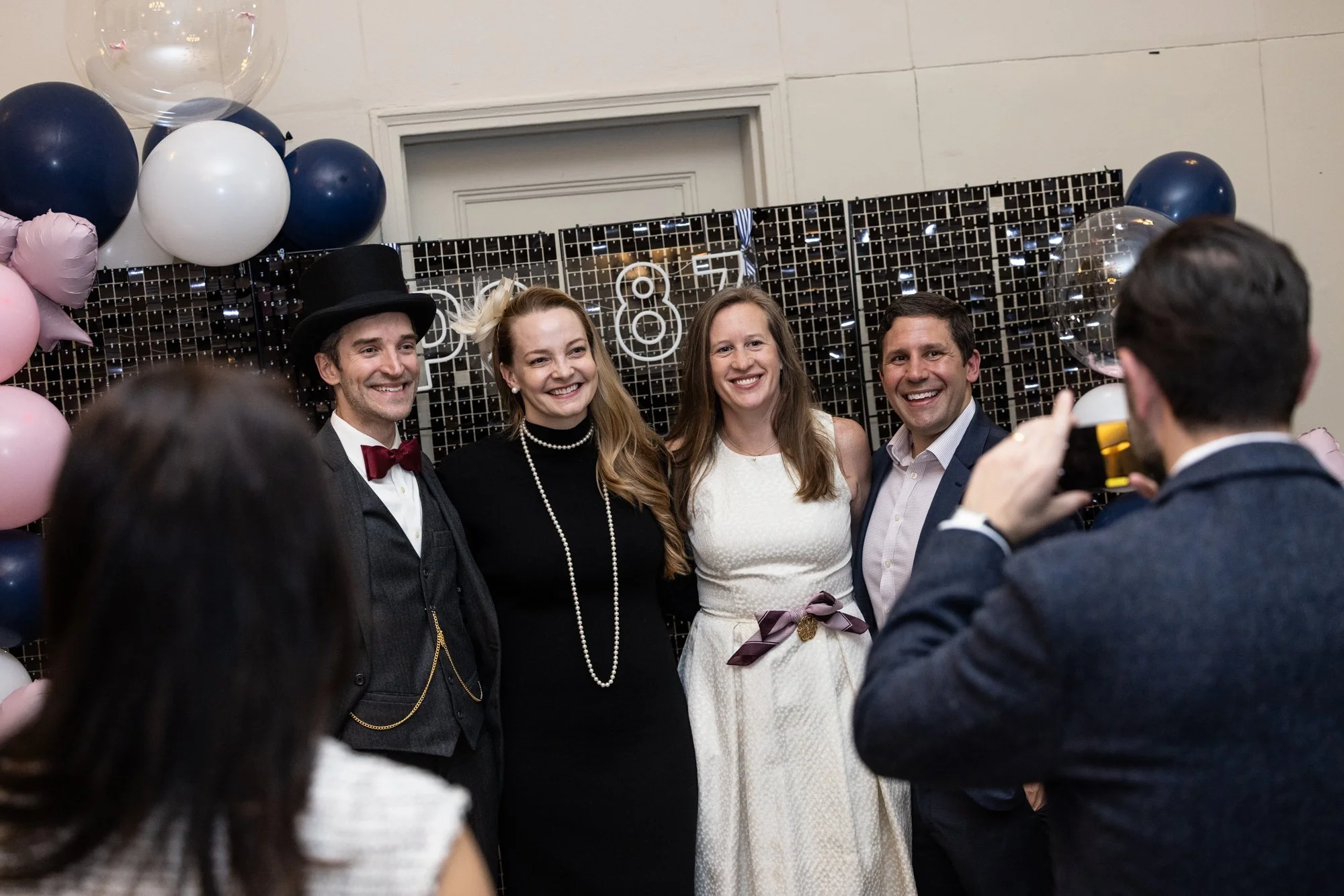Group of four people taking a photo together at a celebration with balloons and a decorated backdrop.
