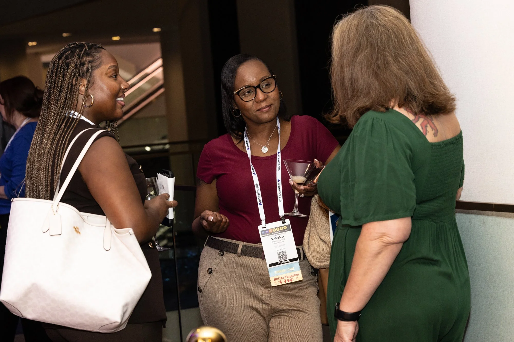 Three women engaged in conversation at a professional event, one holding a wine glass. They are wearing conference badges with one labeled Vanessa. The background is indoors with warm lighting.