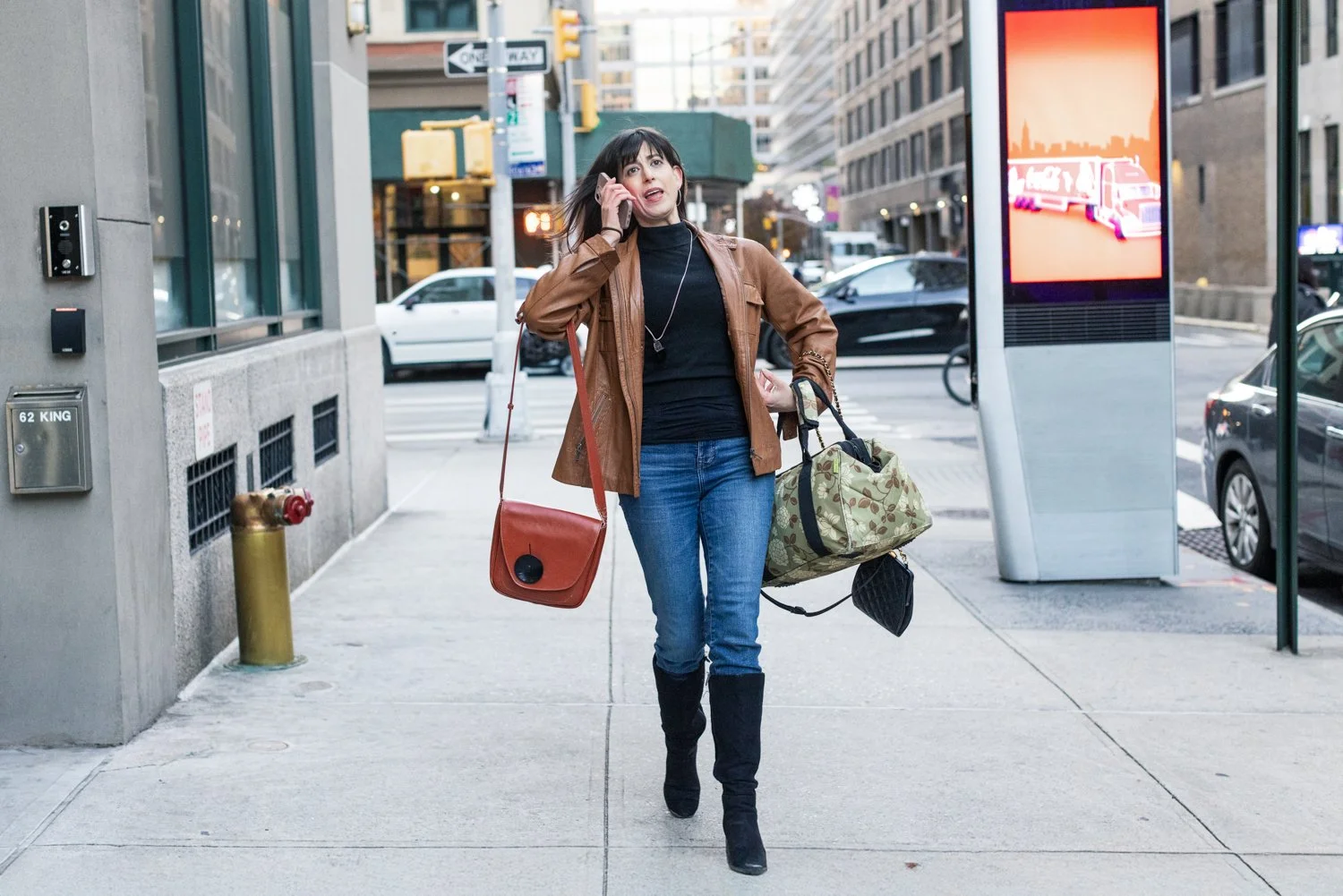 A woman walking on city sidewalk talking on her cellphone, carrying a red shoulder bag and a large camouflage duffel bag.