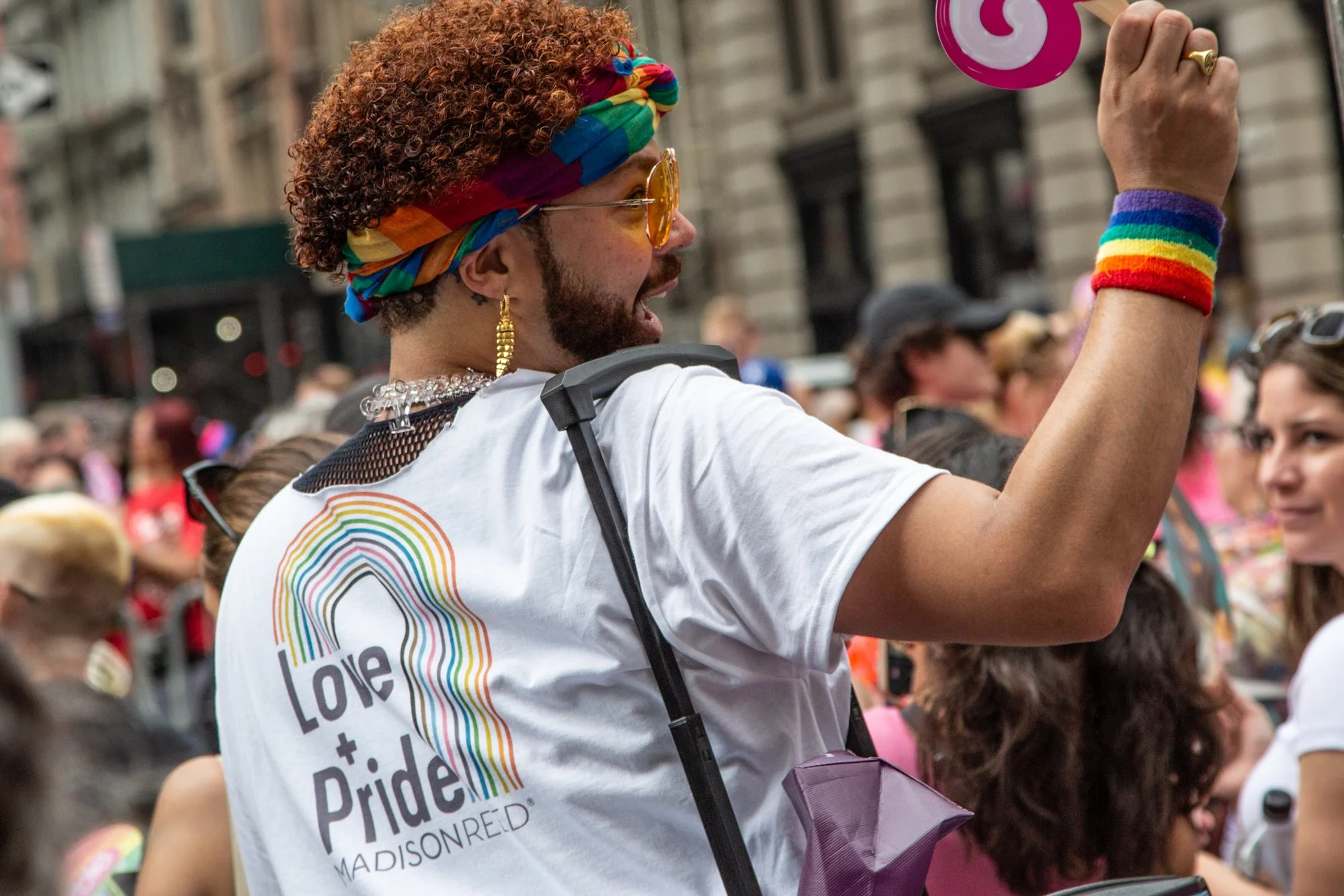 Pride parade participant wearing a rainbow headband, sunglasses, and Love Is Love shirt while raising a hand in a crowd