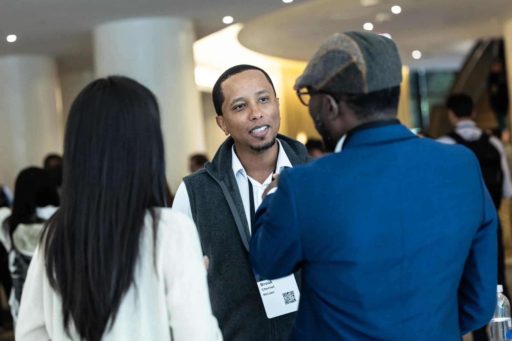Three people in a conversation at a professional event, one man with a name tag, in a modern, well-lit conference hall.