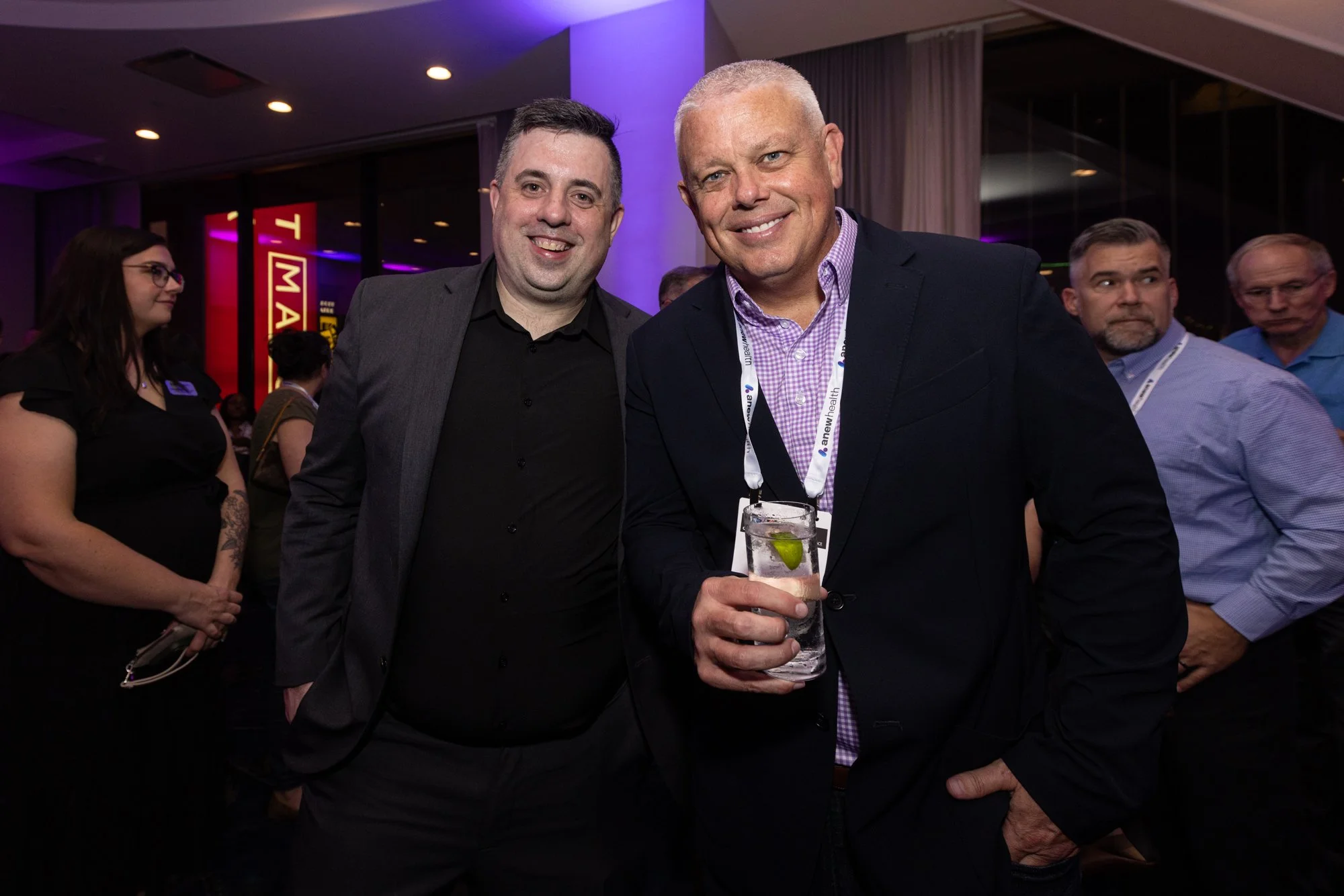 Two men in suits smiling at the camera, one holding a drink with a lime wedge, at an indoor event with purple lighting. Several other people are visible in the background.