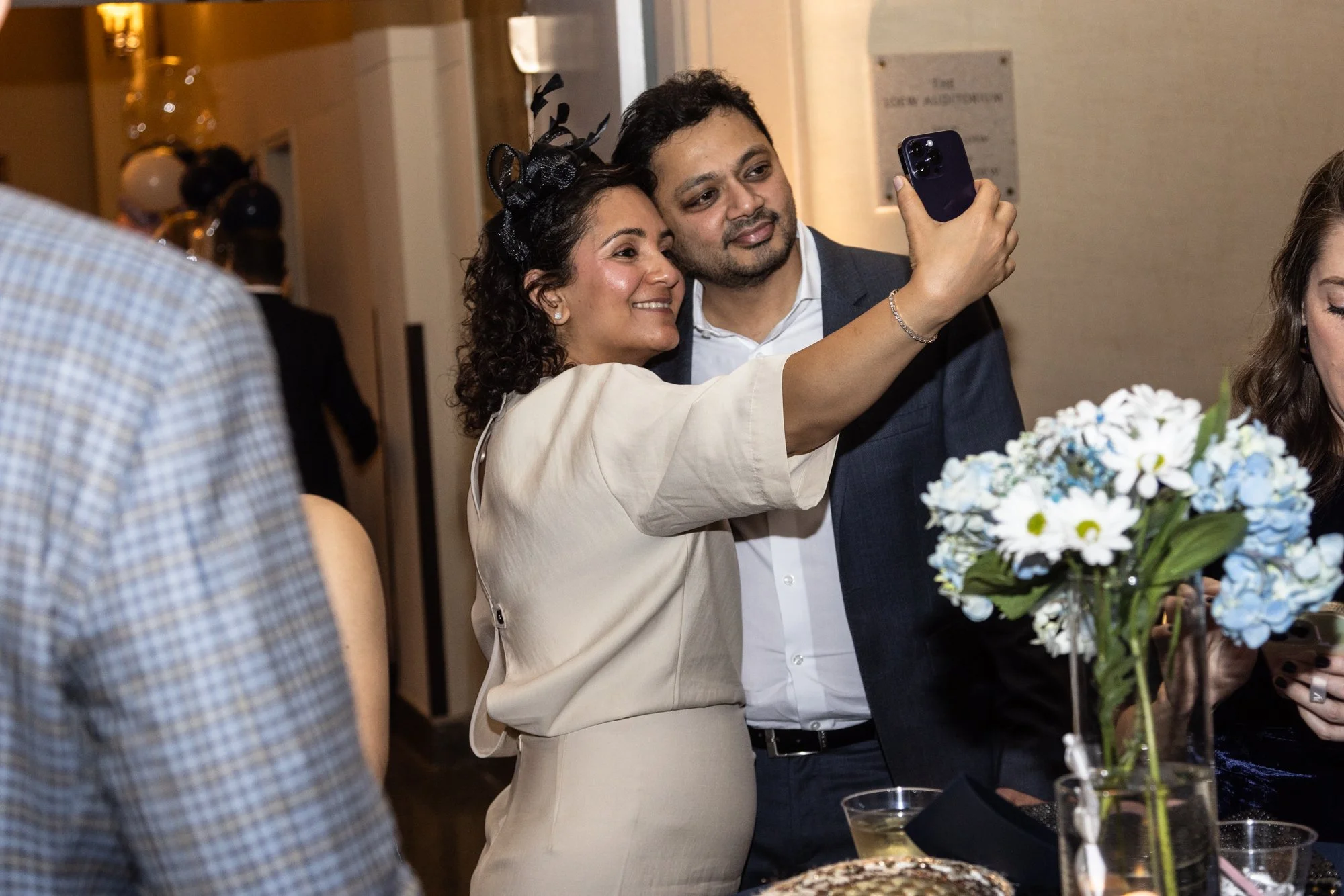 A woman and a man taking a selfie together at a social event. The woman is wearing a beige blazer and a black fascinator, while the man is in a suit. There are flowers and other people visible in the background.