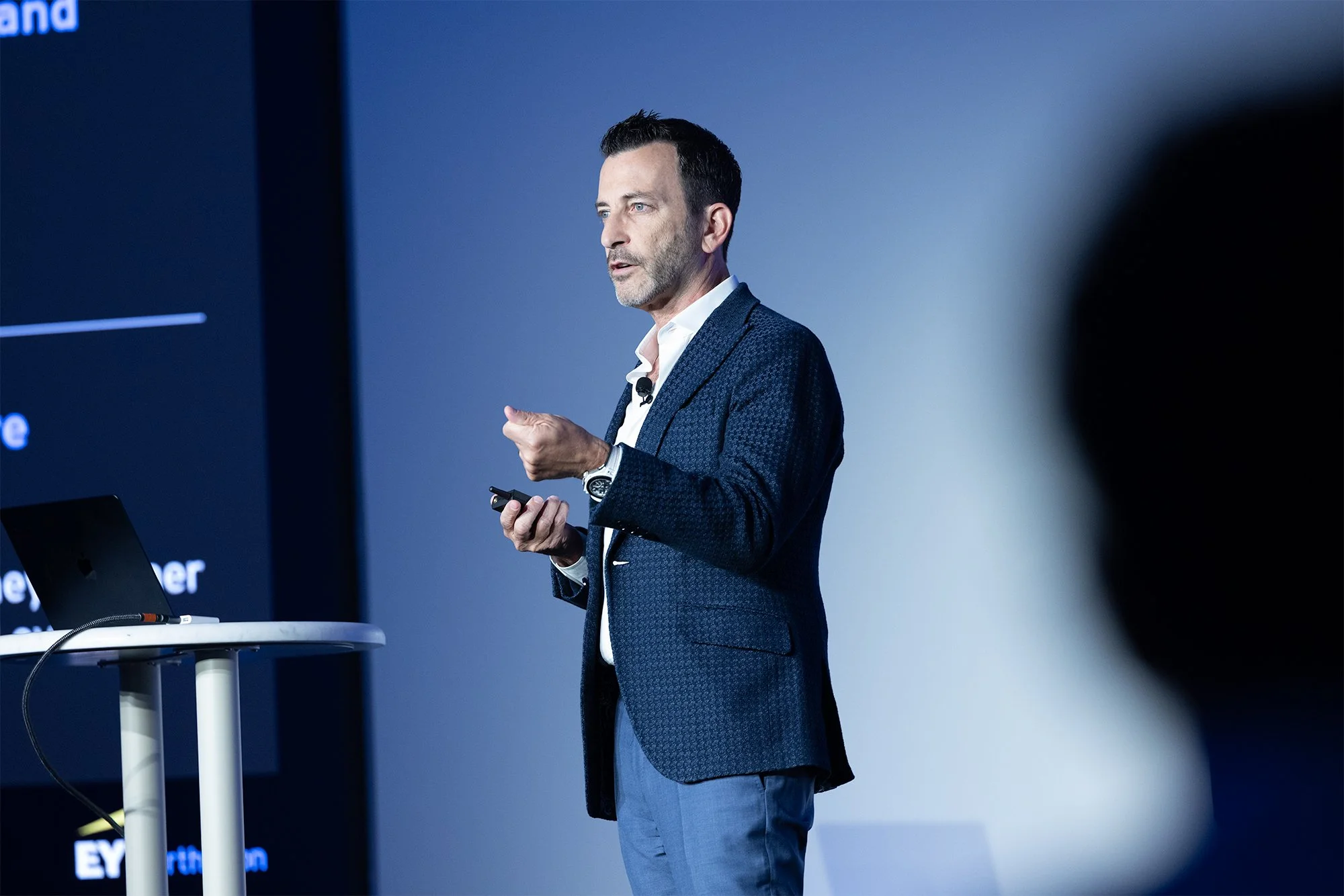A man with dark hair, wearing a blue patterned blazer, white shirt, and jeans, delivering a presentation on stage with a microphone clipped to his shirt.