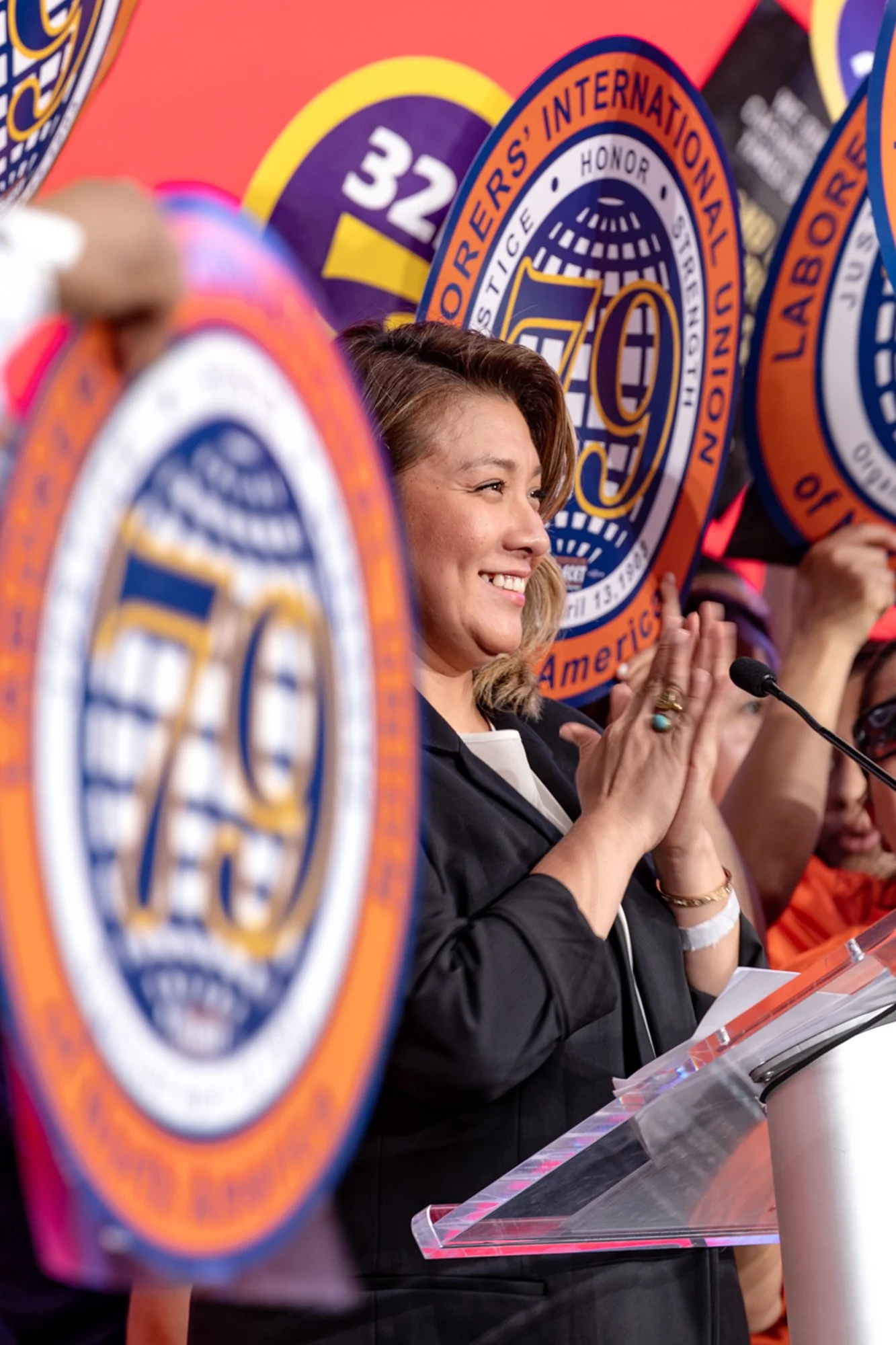A woman with short blond hair smiling and clapping at a conference, surrounded by patches of the FBI and other law enforcement agencies.