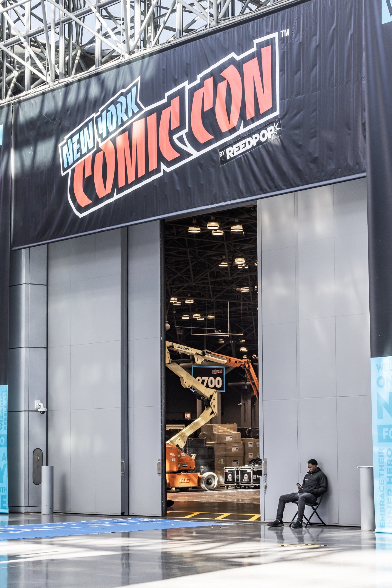 New York Comic Con entrance banner above an exhibition hall doorway with rental lifts, equipment crates, and a seated attendee.