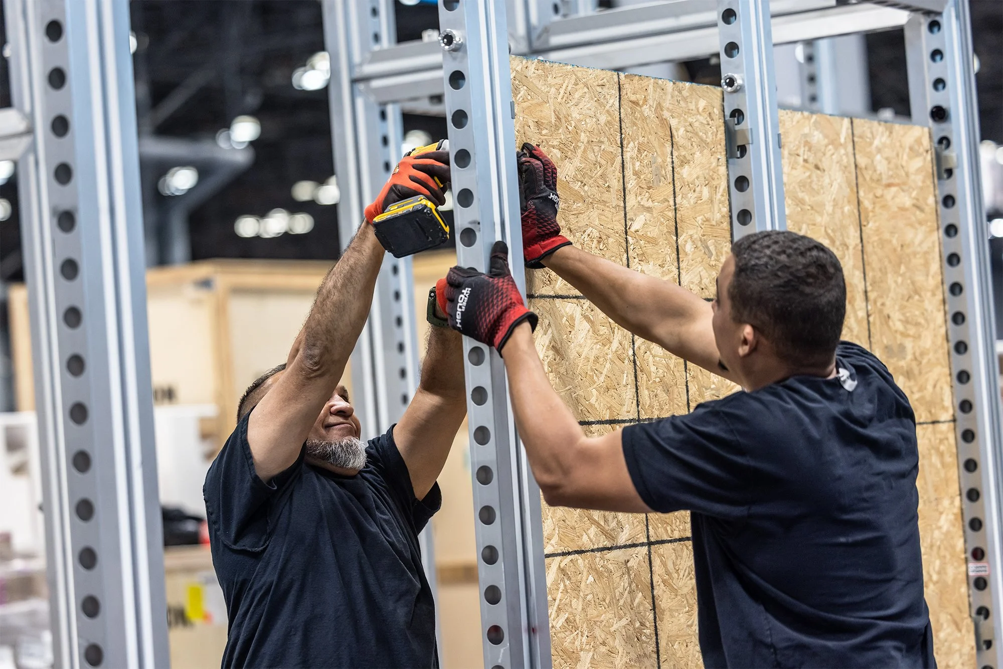 Two construction workers wearing gloves install OSB sheathing within a metal wall framing system.