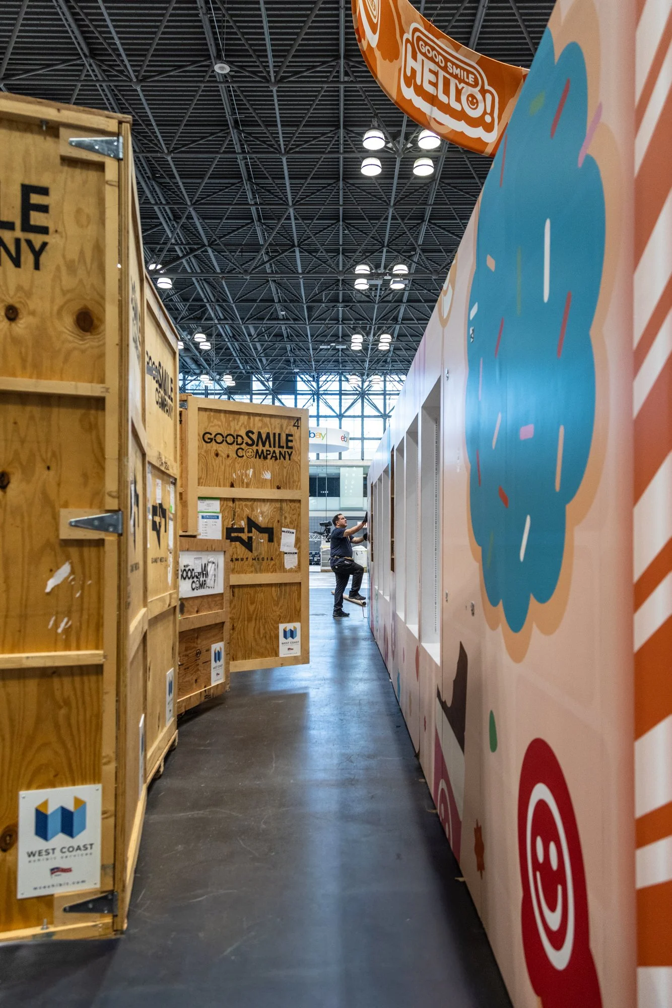 Trade show aisle with Good Smile Company shipping crates, colorful branded booth walls, and a worker assembling the exhibit.