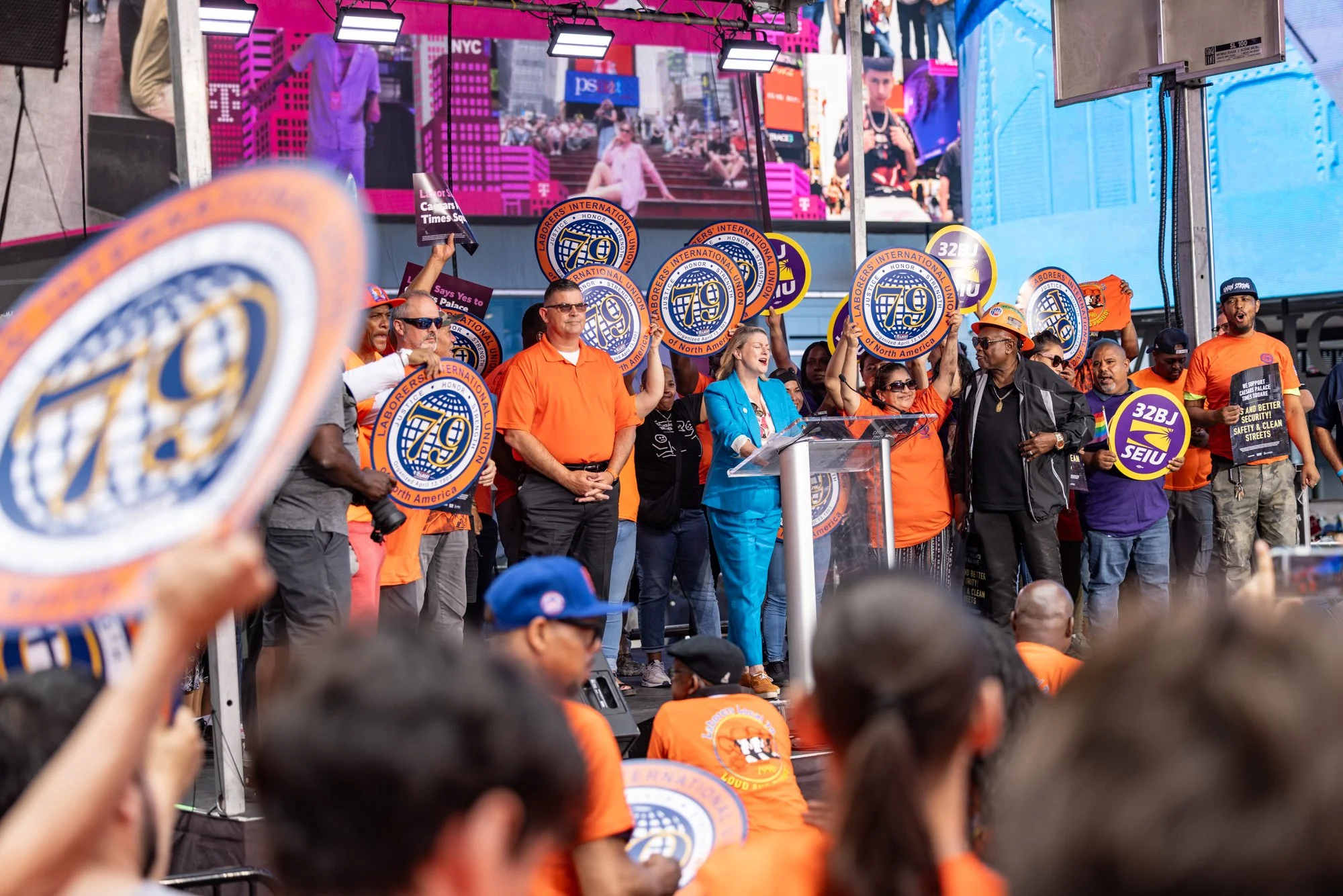 Group of diverse people on a stage at a rally, holding signs that say 'Laborers International Union of North America' and '70,' with a large crowd in front and a large screen displaying the scene.