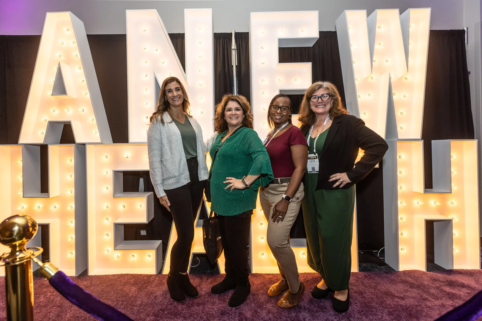 Four women standing side by side in front of large illuminated letters spelling 'LOVE' at an event.