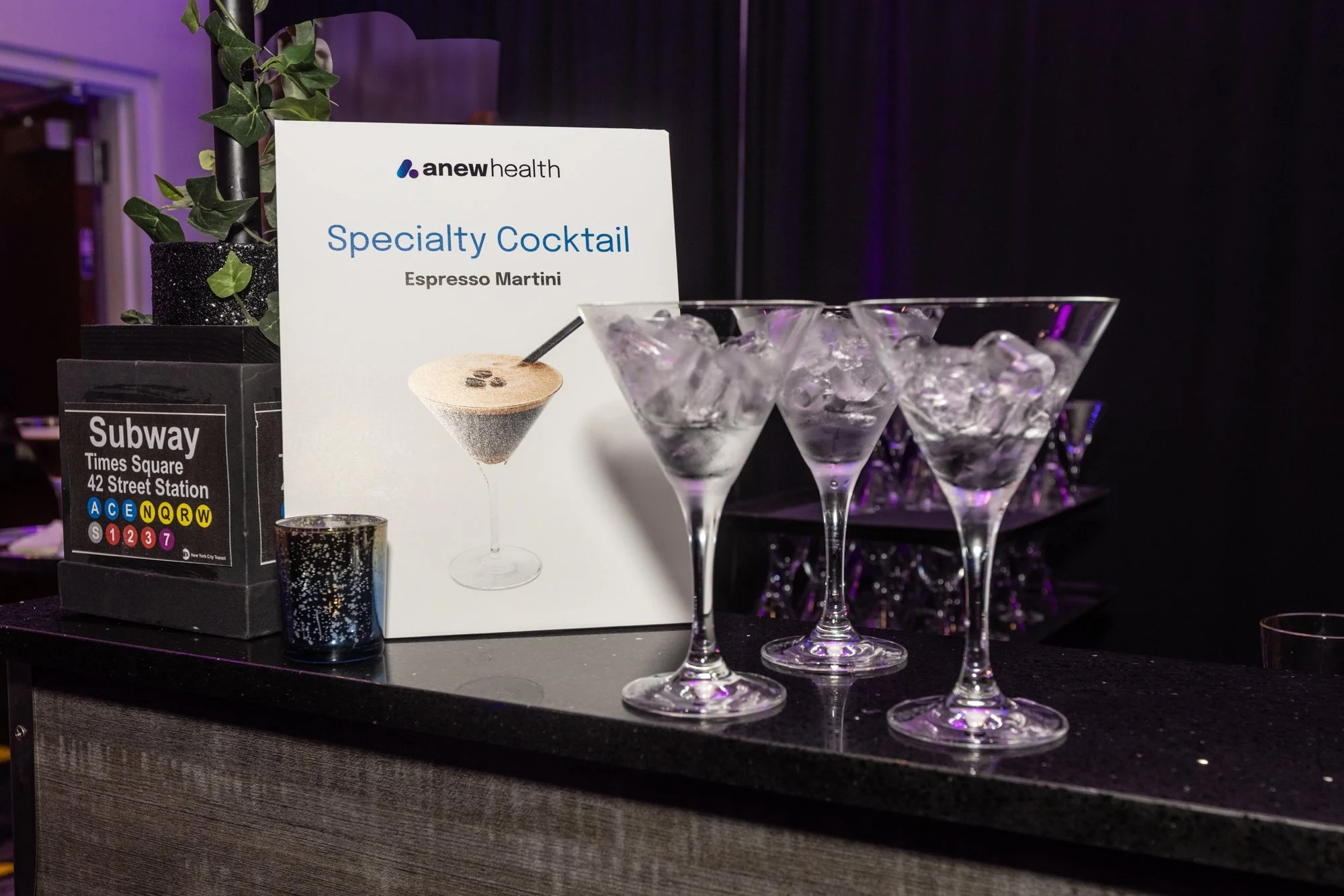 A display at an event shows a sign for a specialty cocktail called Espresso Martini, with a photo of the cocktail. There are glasses filled with ice on the table, and a small potted plant. A subway map and some other objects are partly visible.