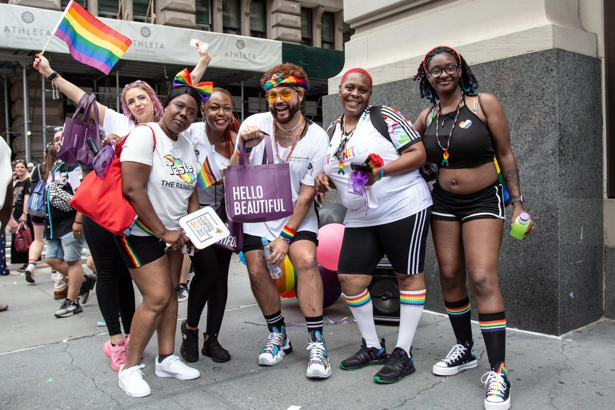 Group of people celebrating Pride with rainbow flags, clothing, and accessories on a city sidewalk.