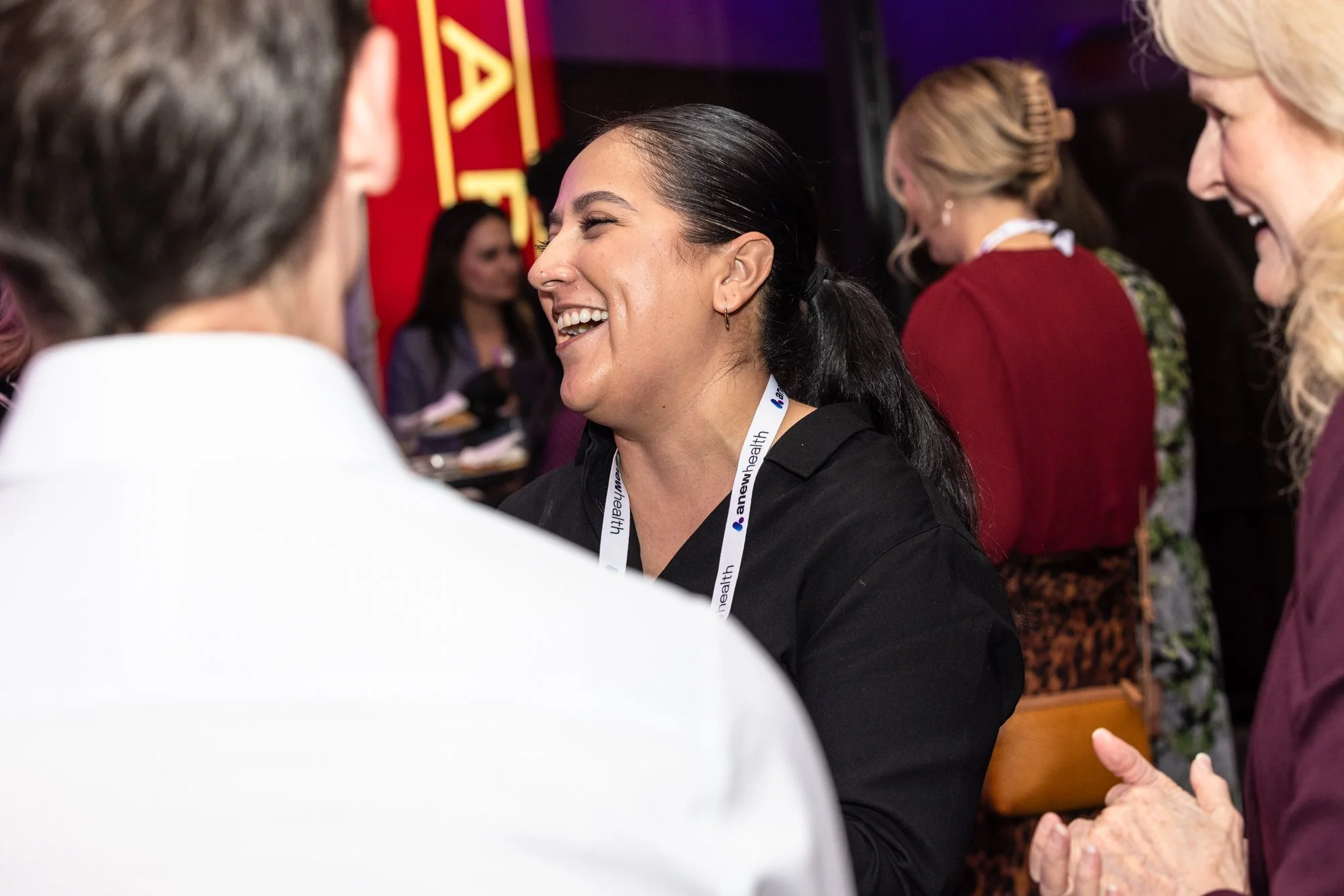 A woman with dark hair tied back and wearing a black shirt laughing and talking with two older women at a social event. The women are engaged in conversation and smiling.